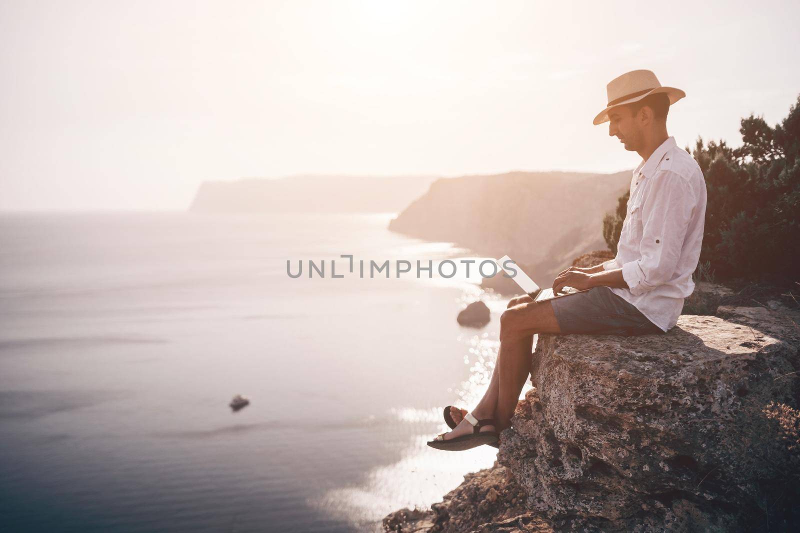 Digital nomad, freelancer. Remote work on vacation. Man in the hat, a businessman with a laptop sits on the rocks by the sea during sunset, makes a business transaction online from a distance. by panophotograph