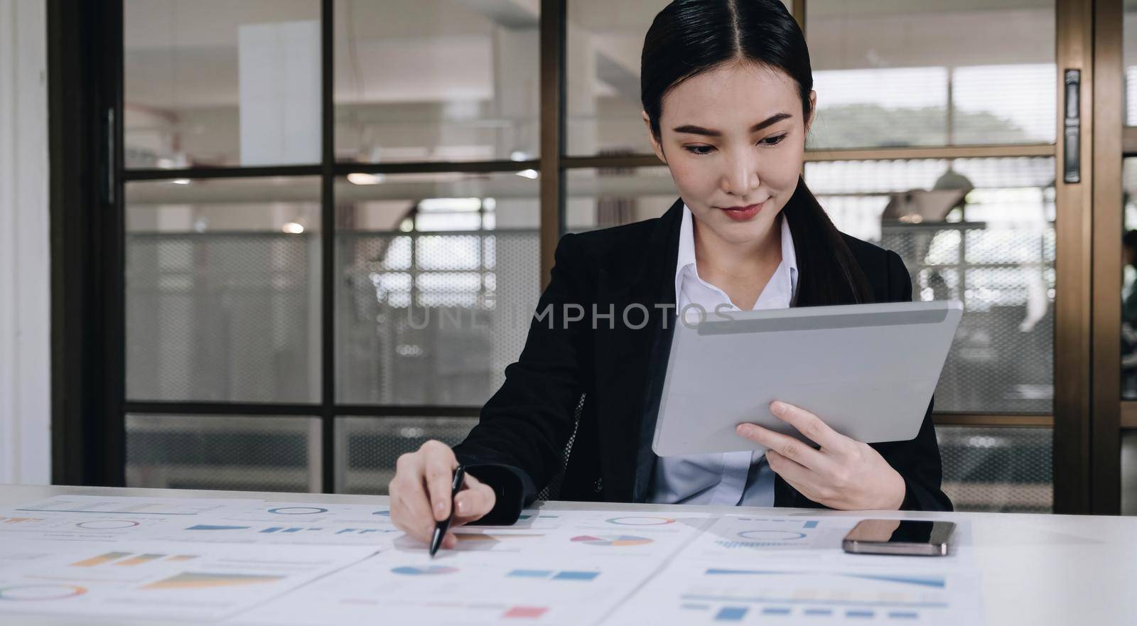 Attractive and charming young Asian businesswoman or financial worker in formal suit using tablet and working with her financial report in the office..