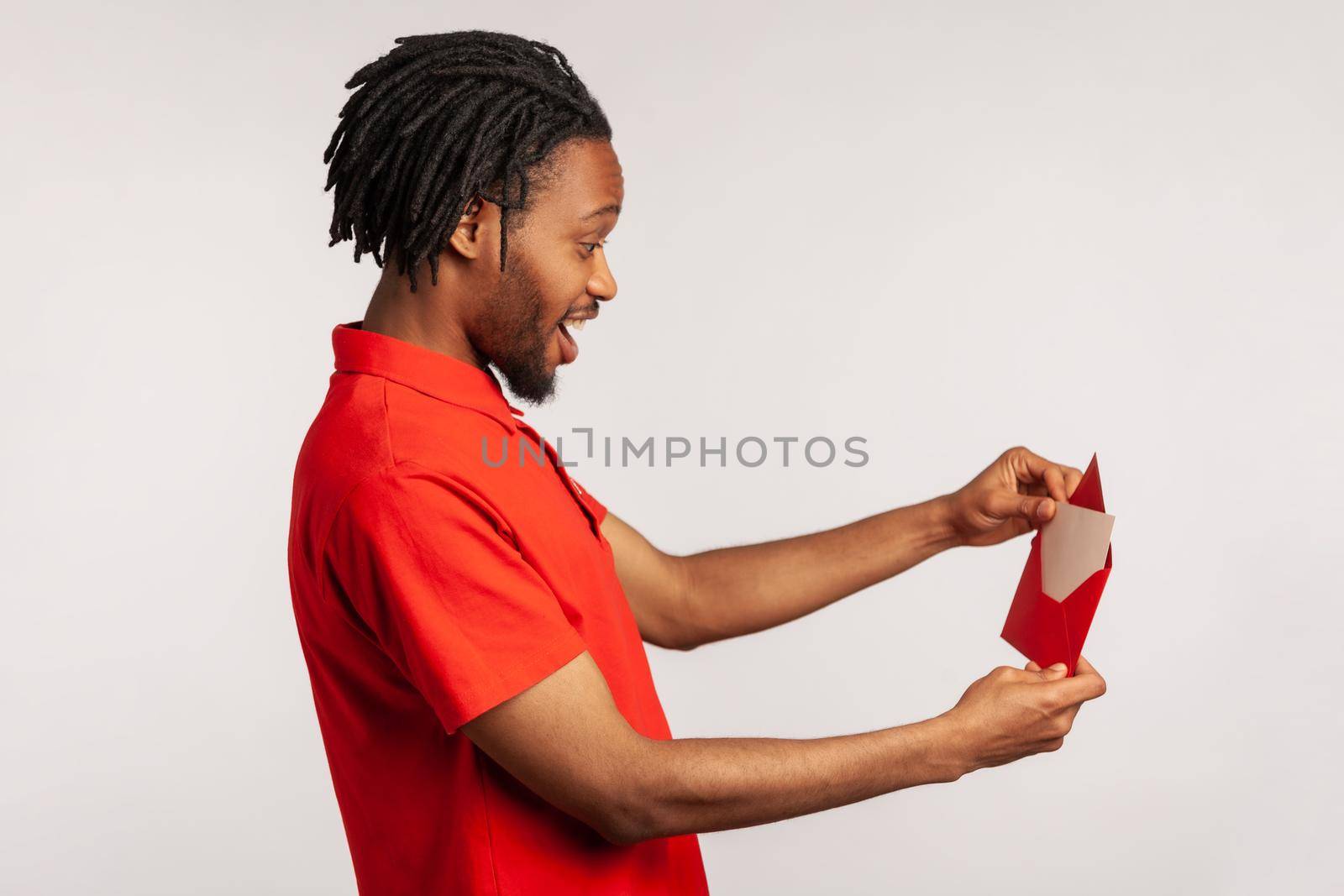 Side view of positive man with dreadlocks wearing red casual style T-shirt, opening letter in red envelope, holding greeting card and feels happy. Indoor studio shot isolated on gray background.