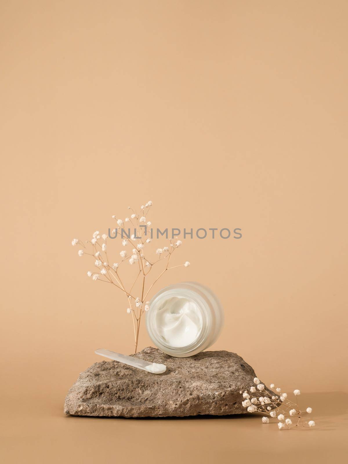 Cosmetic cream or moustirizer on stone over pastel beige background. Open round glass jar with aesthetic swirls face cream and spatula. Copy space. Vertical. Mockup