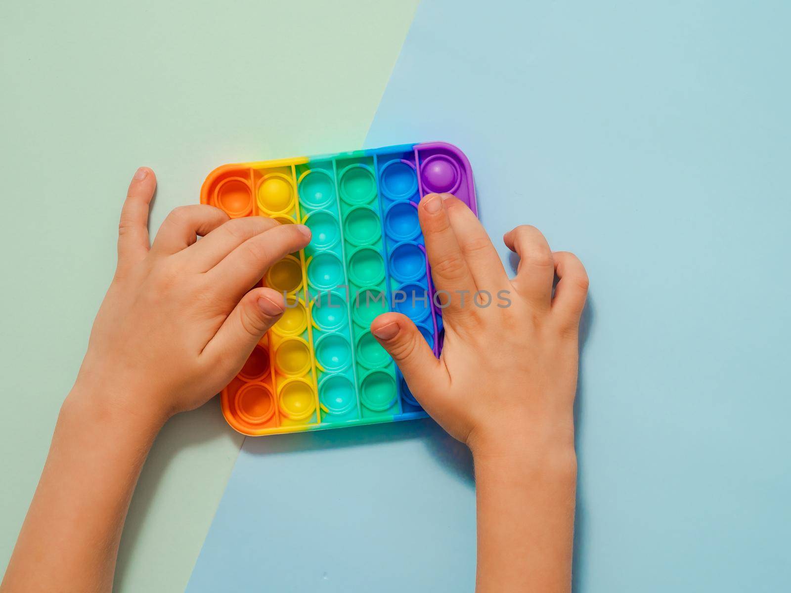 Kid hands play popit toy, top view over two-shadows blue background. Popit on blue background, copy space. Popular silicone colorful antistress pop it toy for child. Top view from above. Simple dimple