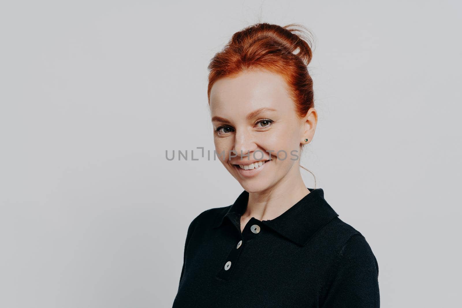 Close up portrait of young beautiful redhead female with natural makeup and beaming smile looking at camera, posing isolated over grey background, attractive female with red hair wearing black t-shirt