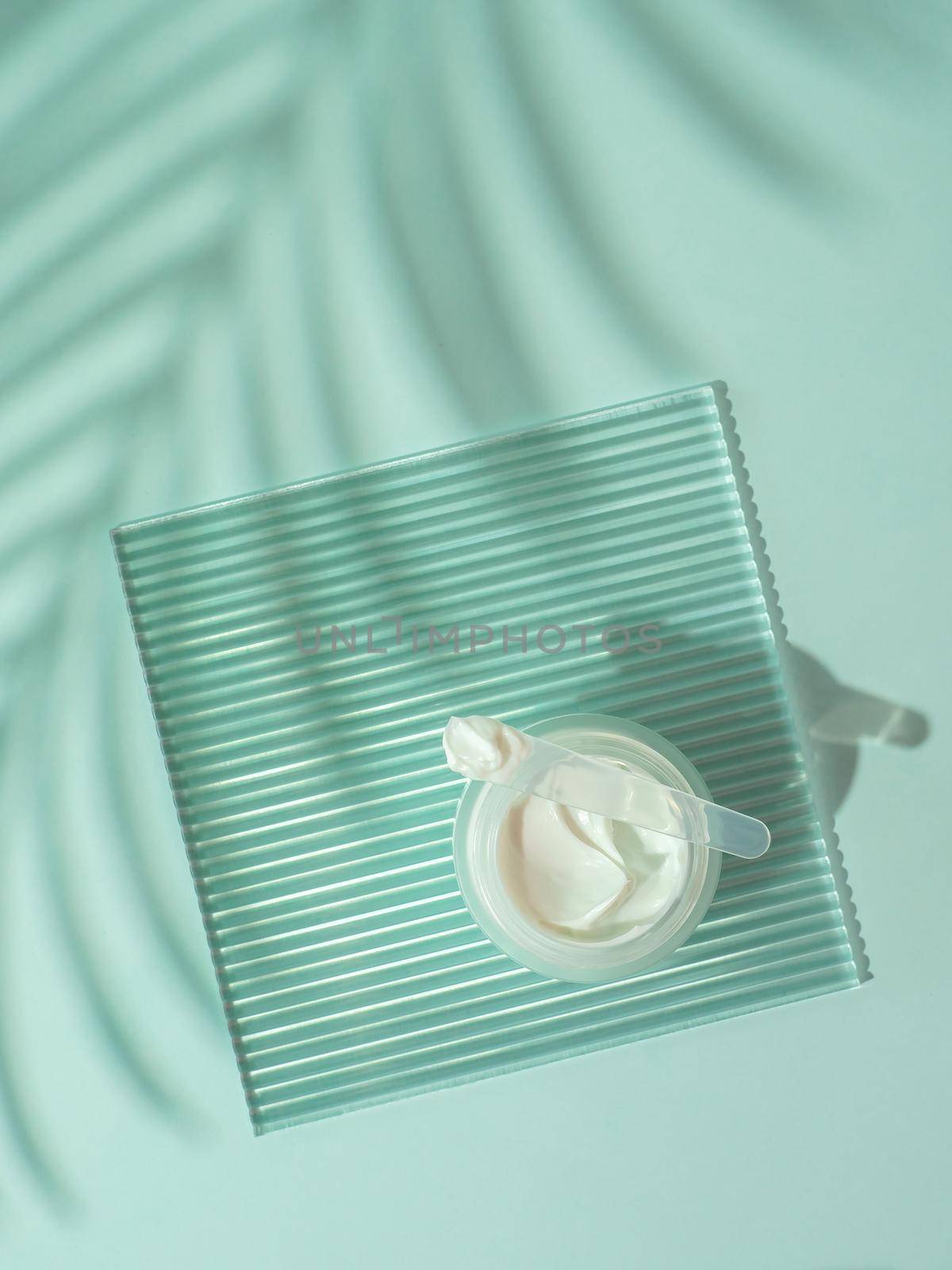 Cosmetic cream moustirizer, transparent ribbed acrylic plate, tropical palm leaf shadows on blue background. Open round glass jar,aesthetic swirls cream,spatula. Top view flatlay. Copy space. Vertical