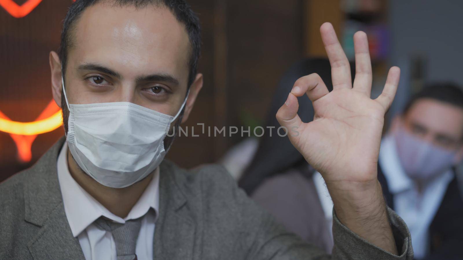 Okey Gesture From Serious Business Man In Protective Mask, Middle Eastern Man Working During Epidemic Of Coronavirus, With Colleagues On Blurred Back, He Looks At Camera In Modern Office