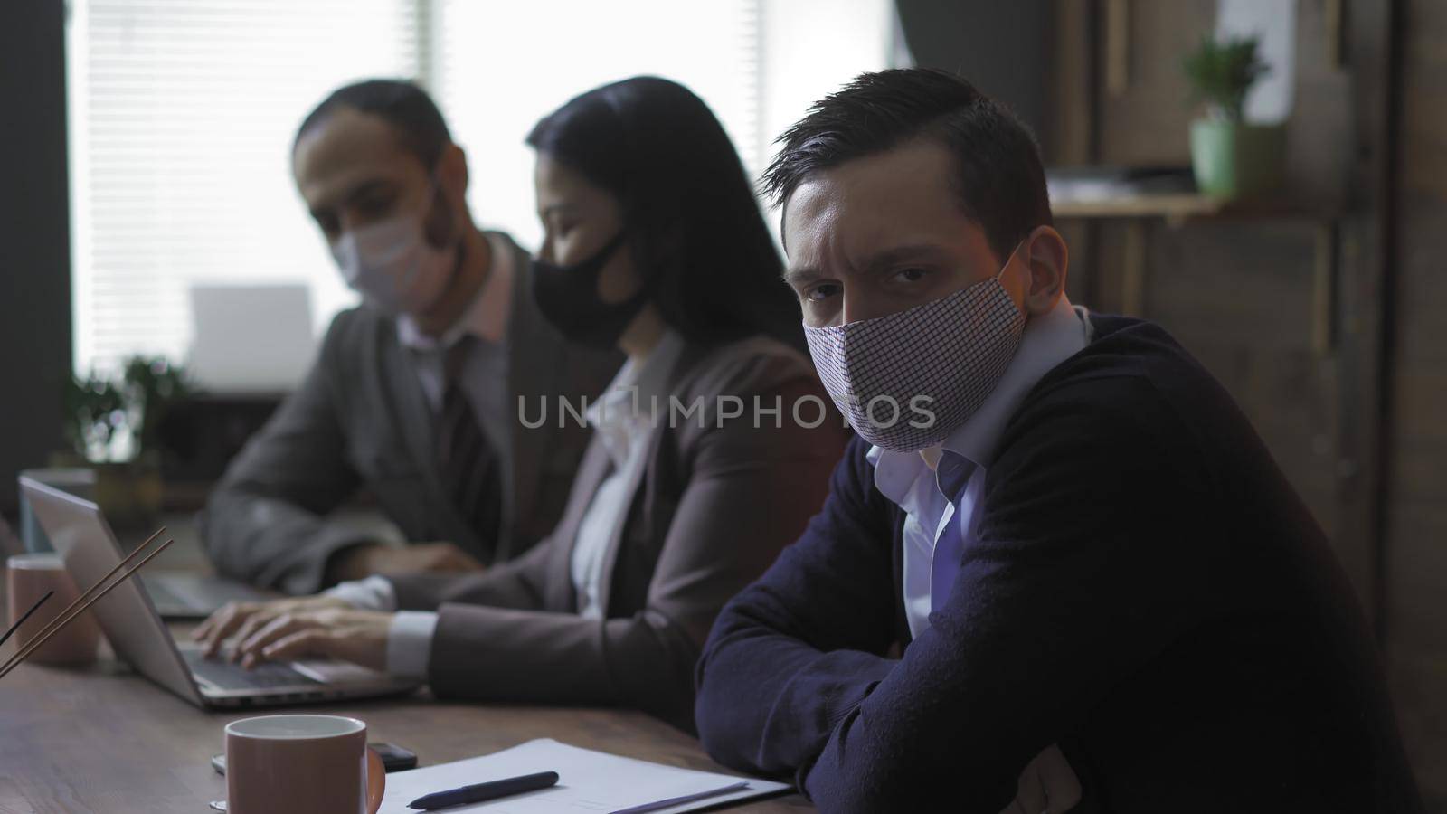 Sad Colleges In Protective Mask Sitting In Office At Meeting During Quarantine From Coronavirus Epidemic, Focus On Young Caucasian Man In Foreground Looking At Camera