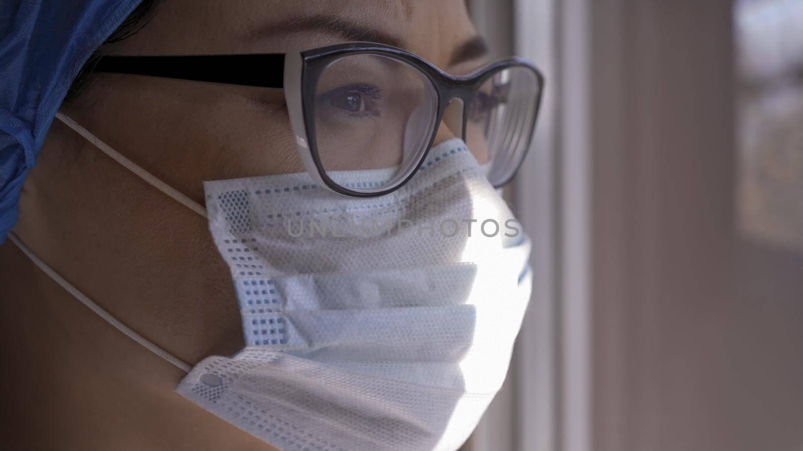 Profile View Of Female Medic Looks At Window In Sunny Day, Portrait Of Sad Young Woman In Protective Mask Forced To Be Isolated During Coronavirus Pandemic, Copy Space At Right Side