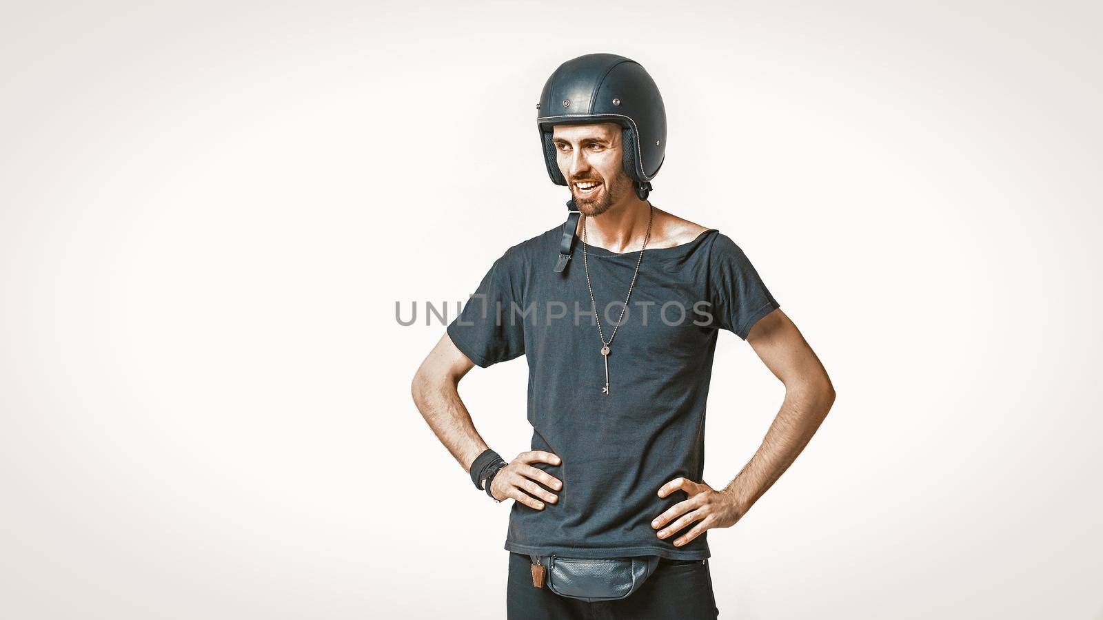 Cute Motorbiker In Protective Helmet Standing Akimbo, Male Portrait Of Caucasian Man In Black Sports Clothing Looks At Left Side Toothy Smiling, Isolated On White, Copy Space At Left Side