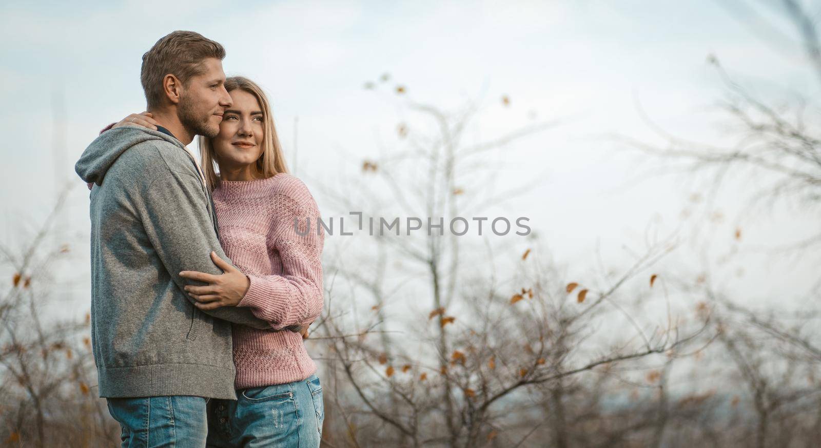 Loving Couple Is Standing In An Embrace In Nature, Young Woman And Man Are Hugging While Admiring The Sunset Against Sky And Autumn Trees, Copy Space At Right Side