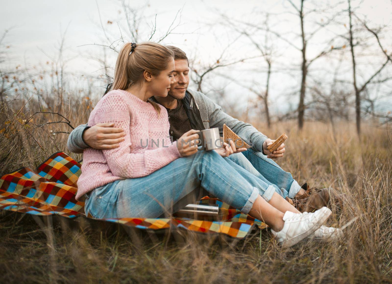 Cheerful Couple Has Romantic Picnic In Nature, Young Man And Woman Eat Snacks And Drink Hot Drink From Cups While Sitting On Picnic Blanket, Caucasian Man Gently Hugs His Woman In Nature
