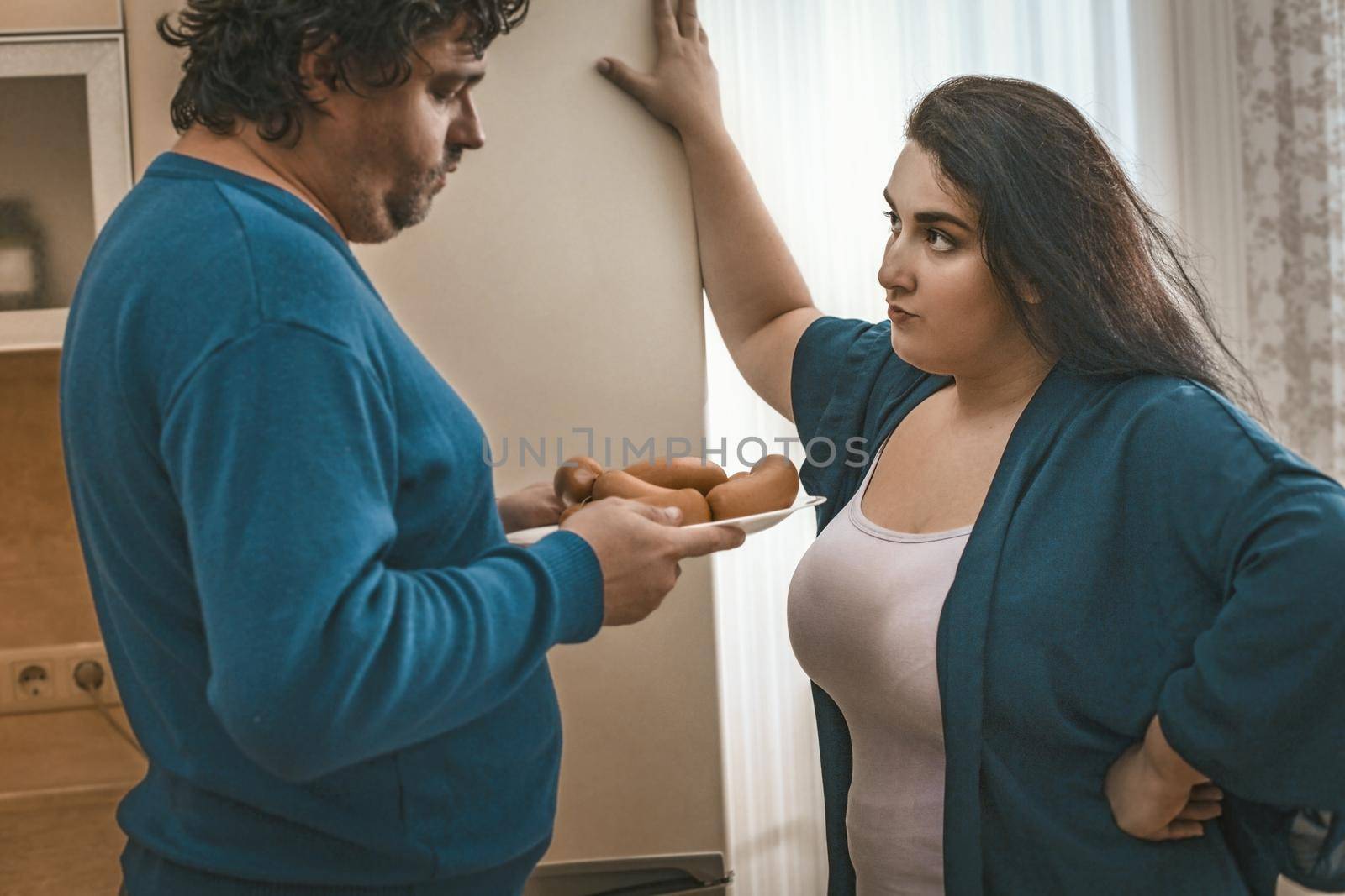 Bodipositive Wife Found That Her Husband Had Violated Diet, Wife Looks Strictly At Her Big Husband Who Is Holding Plate Of Sausages, Couple Standing In Home Kitchen Against Background Of Refrigerator