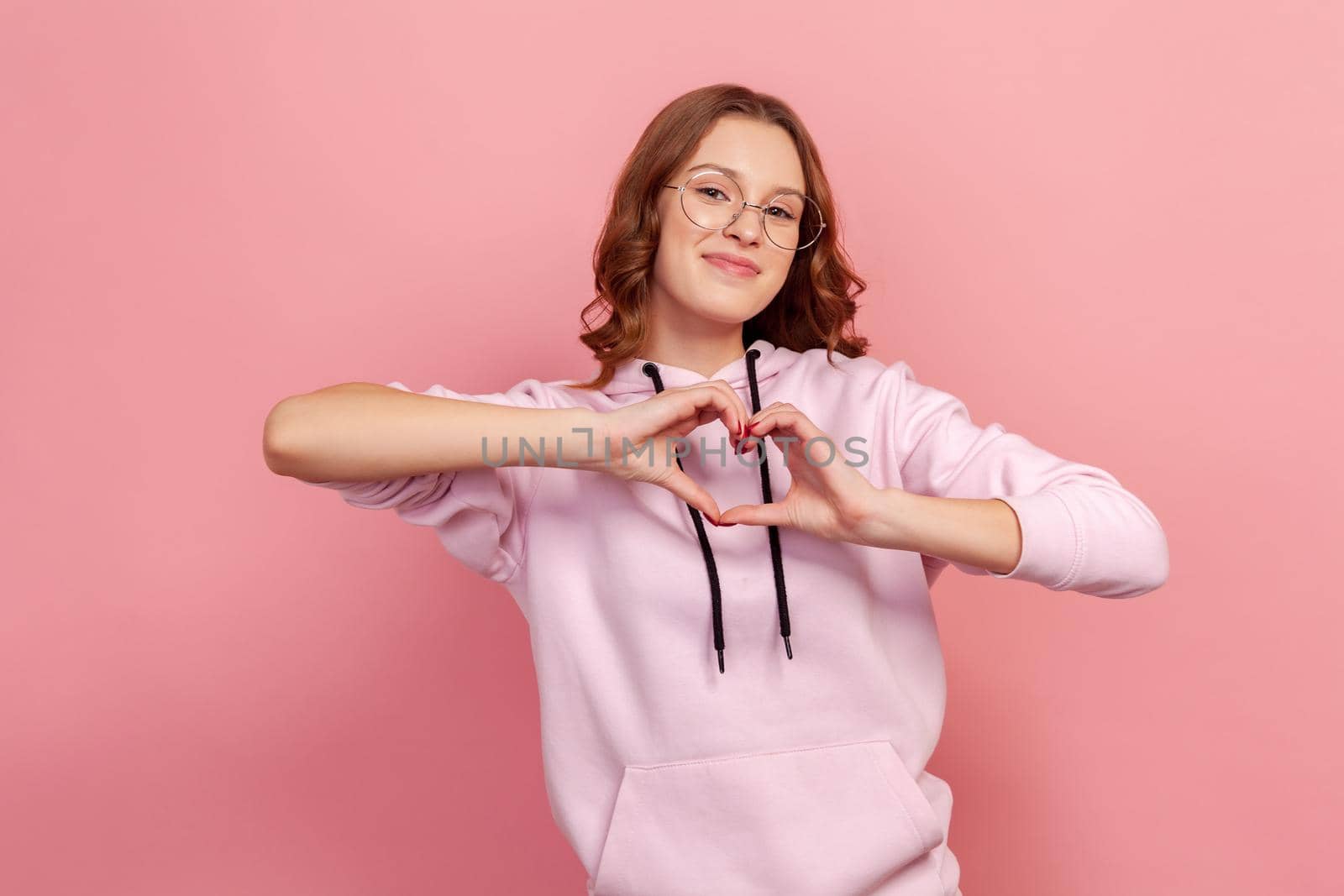 Portrait of cute smiling teen girl in eyeglasses and hoodie making heart shape with hands, expressing love, friendship and care, romance in adolescence. Indoor studio shot isolated on pink background