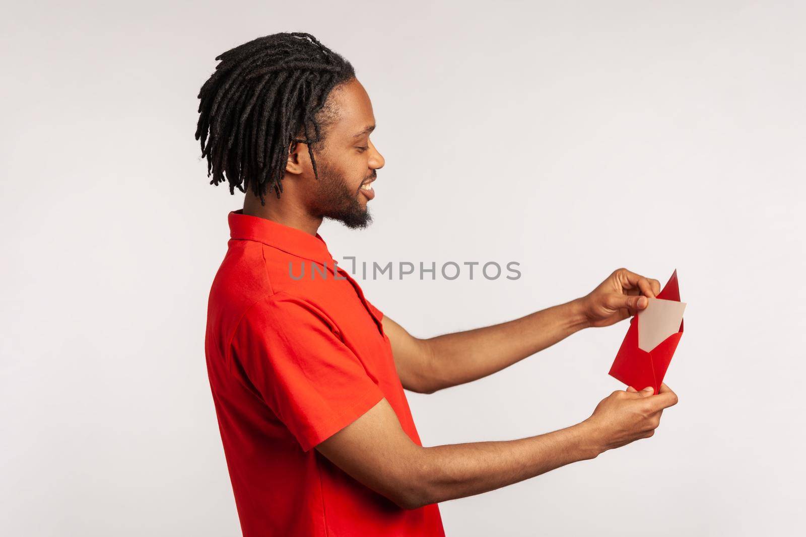 Profile of man with dreadlocks wearing red casual style T-shirt, reading letter or greeting card, holding envelope, smiling and rejoicing pleasant news. Indoor studio shot isolated on gray background.