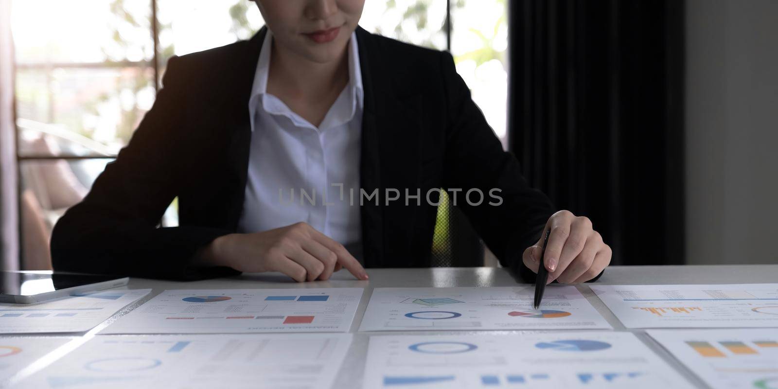 Young Asian businesswoman analyzing report pointing at graph holding a pen using a tablet laptop placed at the office..