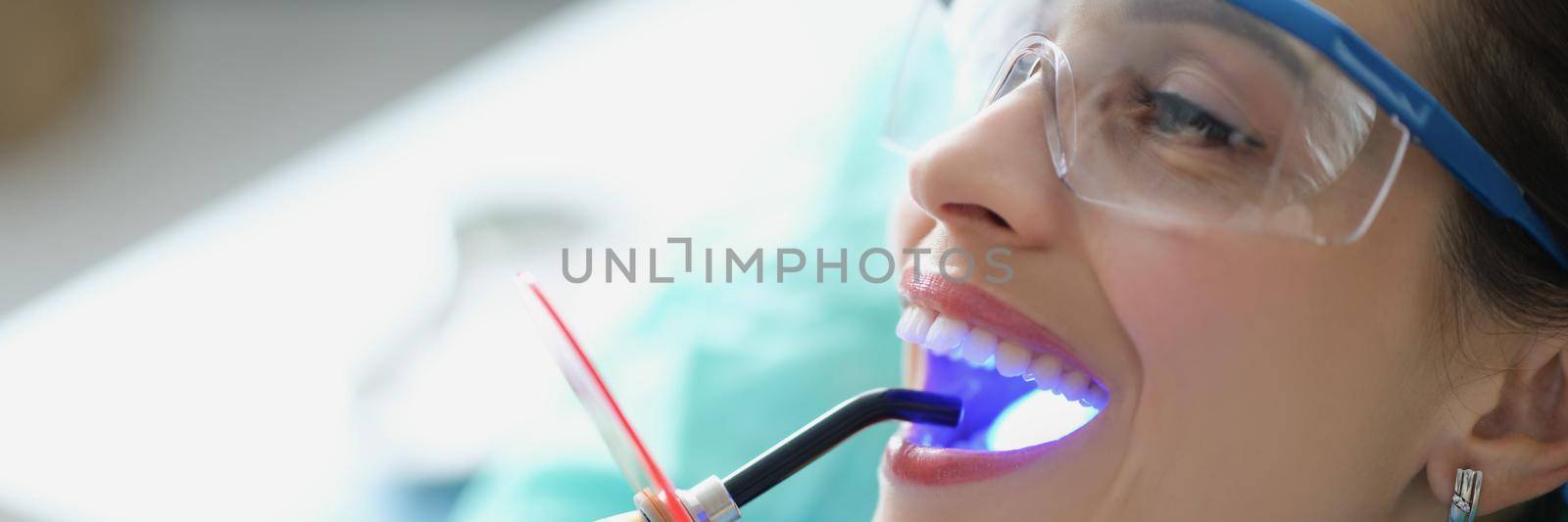 Portrait of female in stomatology cabinet seat getting whitening procedure, open mouth widely. Dentist provide full teeth care for patient. Health concept