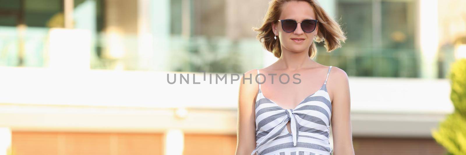 Portrait of beautiful lonely woman walking on hotel territory on sunny summer day. Blonde female in light cute dress on resort background. Holiday concept