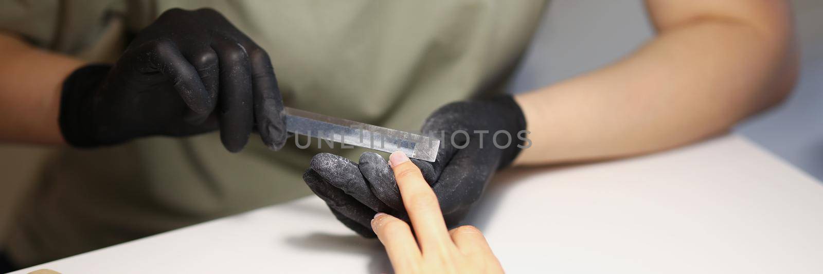 Close-up of nail master using nail file tool to create perfect shape for clients nails. Worker wear protective black gloves for work. Beauty studio concept