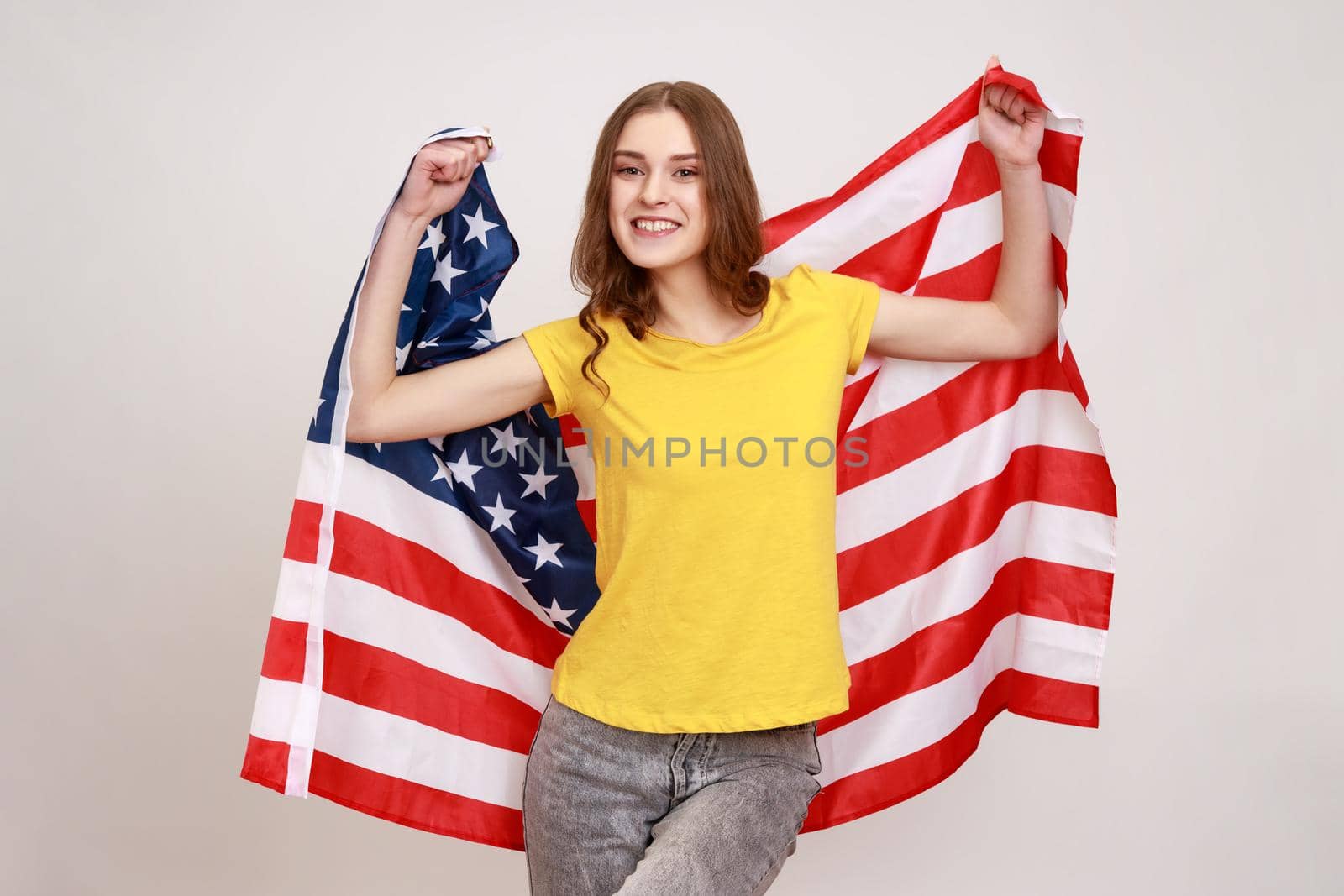 Smiling woman of young age with pleasant appearance and wavy hair, wears casual style yellow T-shirt, holding USA flag and looking at camera with smile. Indoor studio shot isolated on gray background.