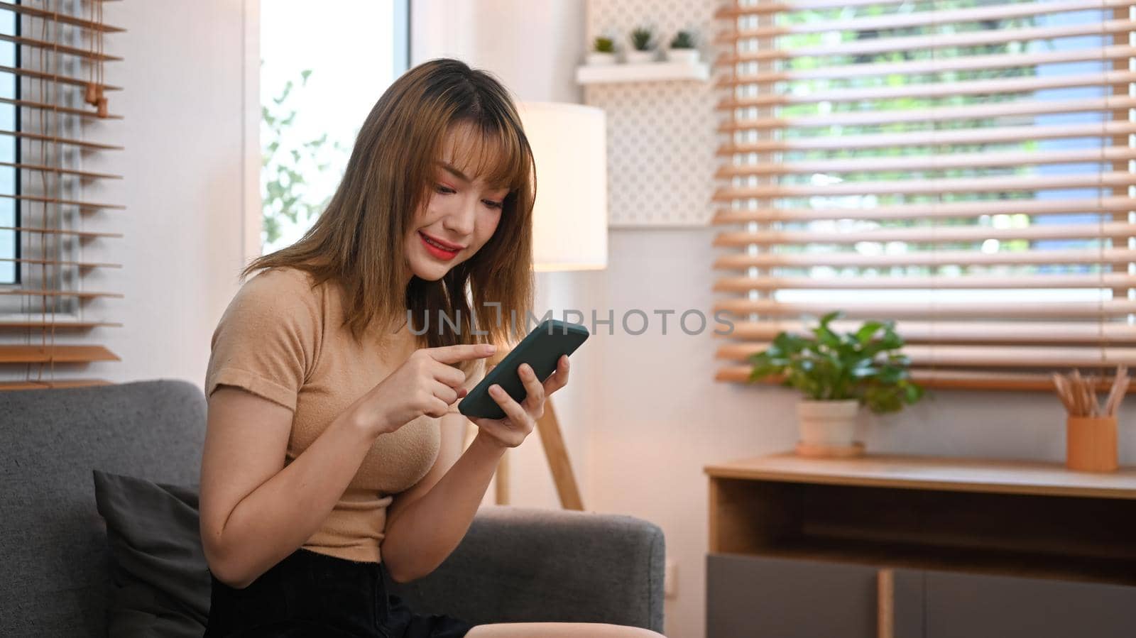 Pretty young woman using mobile phone for chatting with friends while resting in comfortable couch at home.