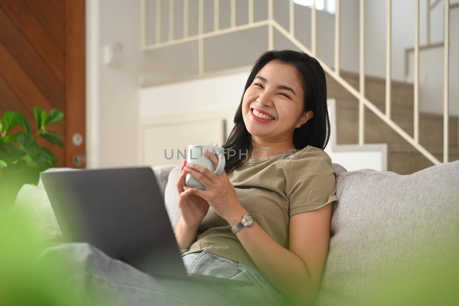 Cheerful asian woman holding coffee cup in her hands and using laptop on coat at home.