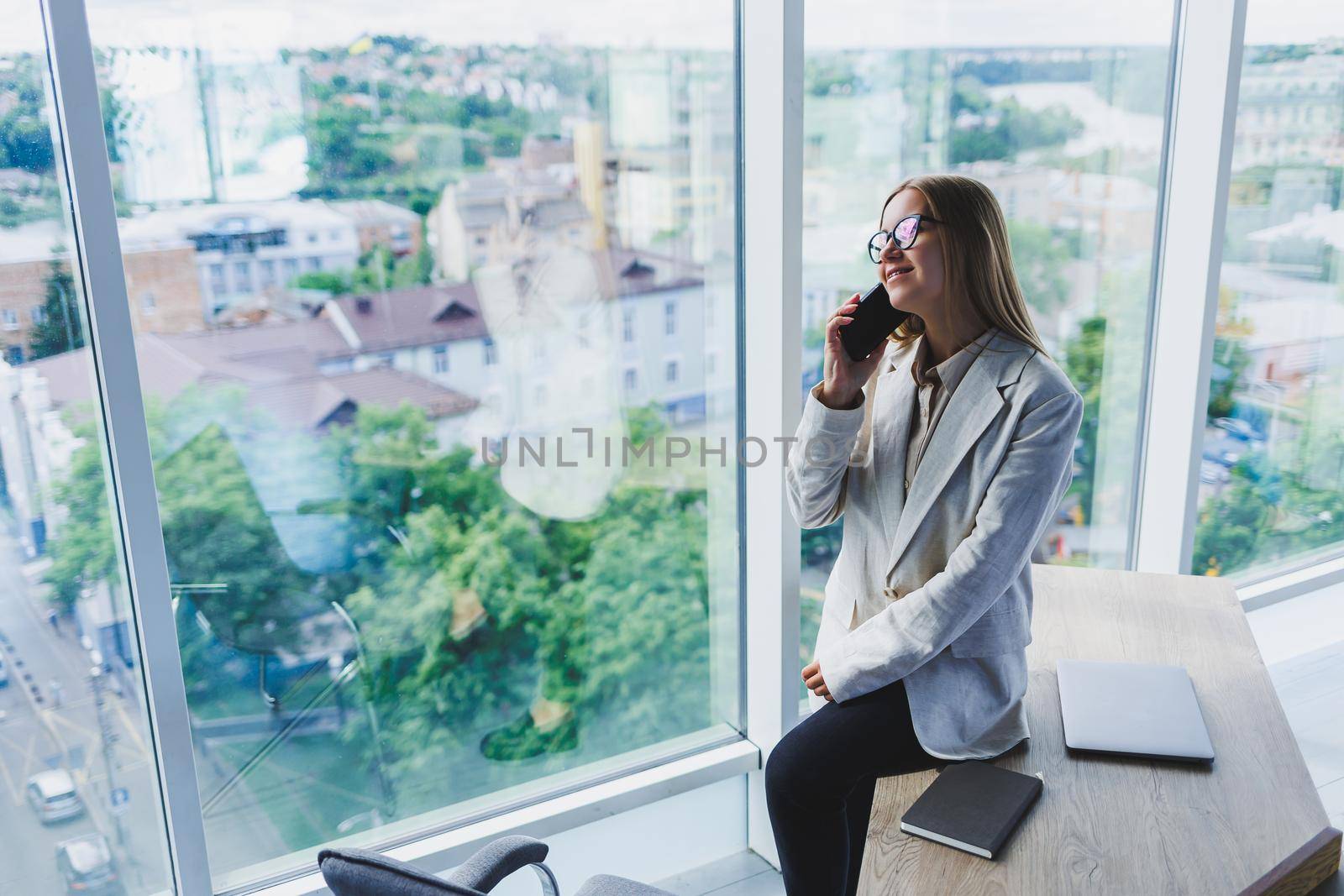 Cheerful young beautiful woman in glasses talking on a mobile phone and using a laptop with a smile while sitting at her workplace. Work in the office