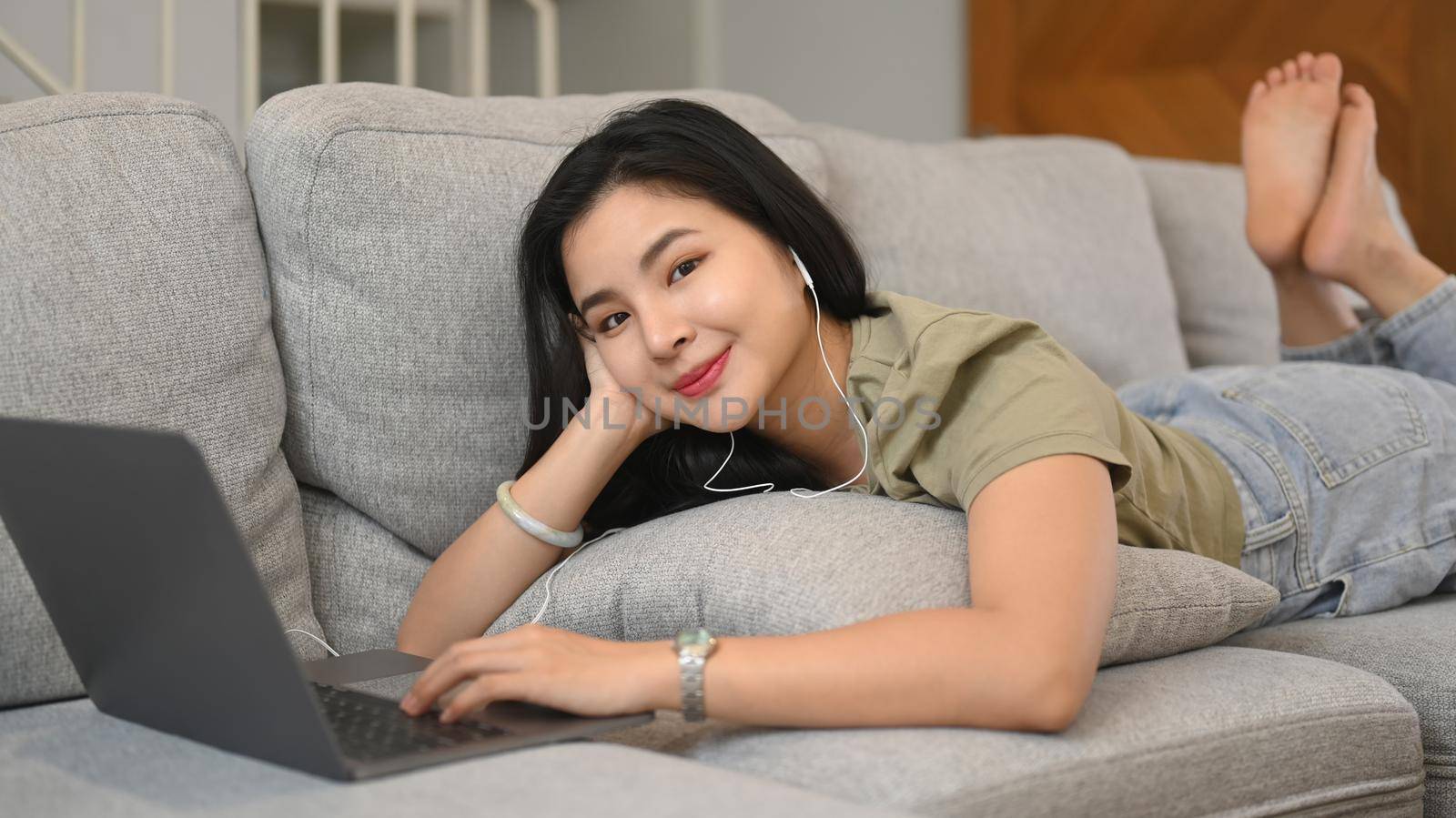 Pretty young asian woman lying on cozy couch and browsing internet via laptop computer by prathanchorruangsak