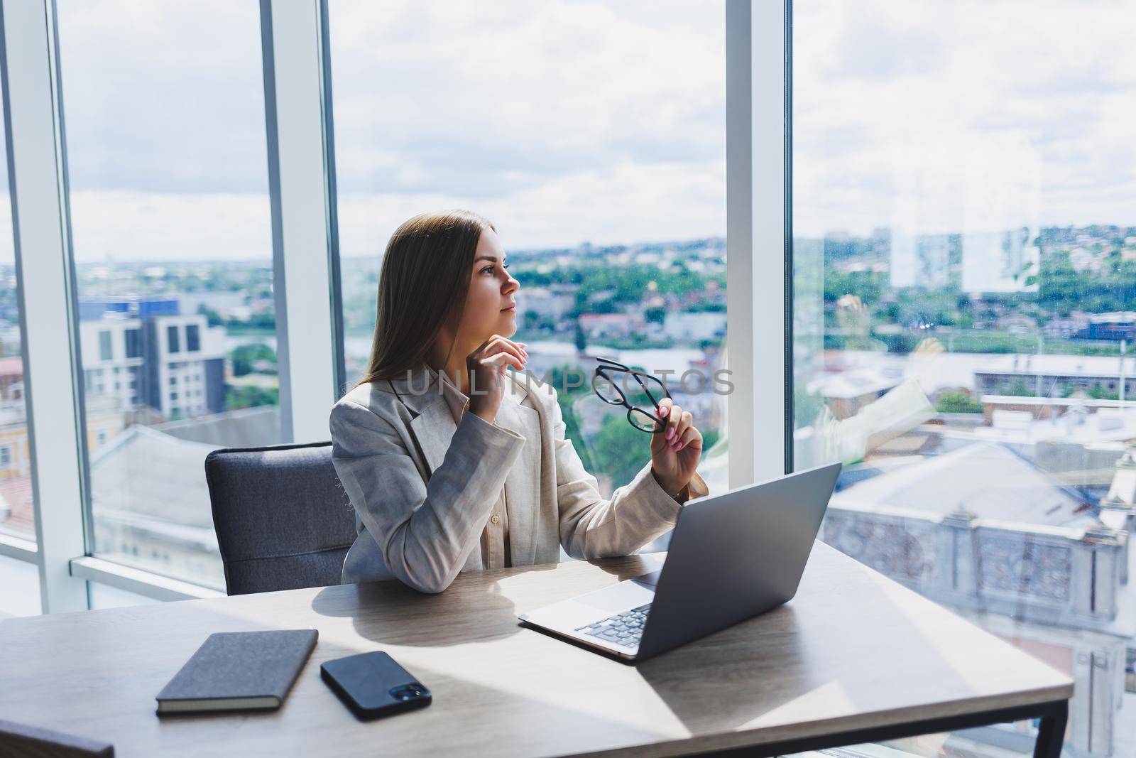 A female head of a company in glasses sits at a laptop in her office with a breathtaking view of the city. Business lady works at a wooden table.