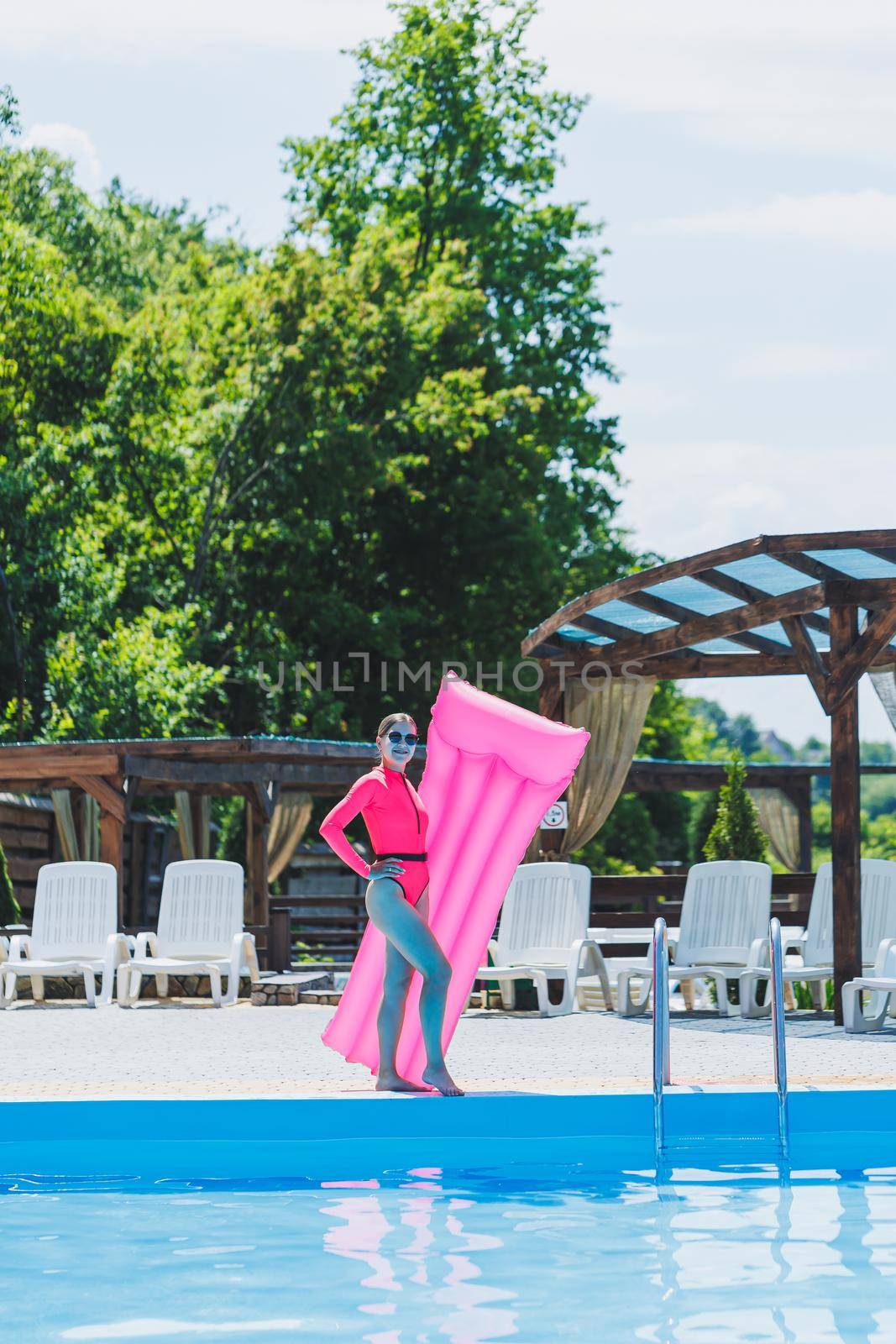 A beautiful woman in a pink swimsuit and with an inflatable mattress stands near a large pool. Summer holidays in a hotel with a swimming pool. Young girl in swimsuit and sunglasses