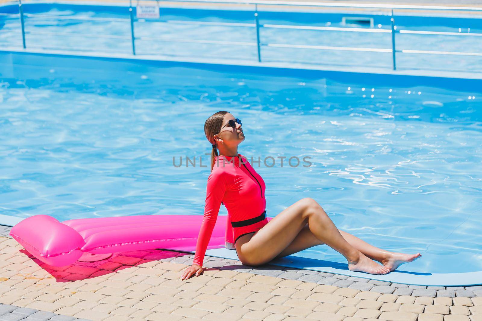 Pretty woman in a bright pink swimsuit near the pool on vacation, she is wearing sunglasses. The concept of summer holidays in the hotel by the pool. Inflatable pink mattress on the background