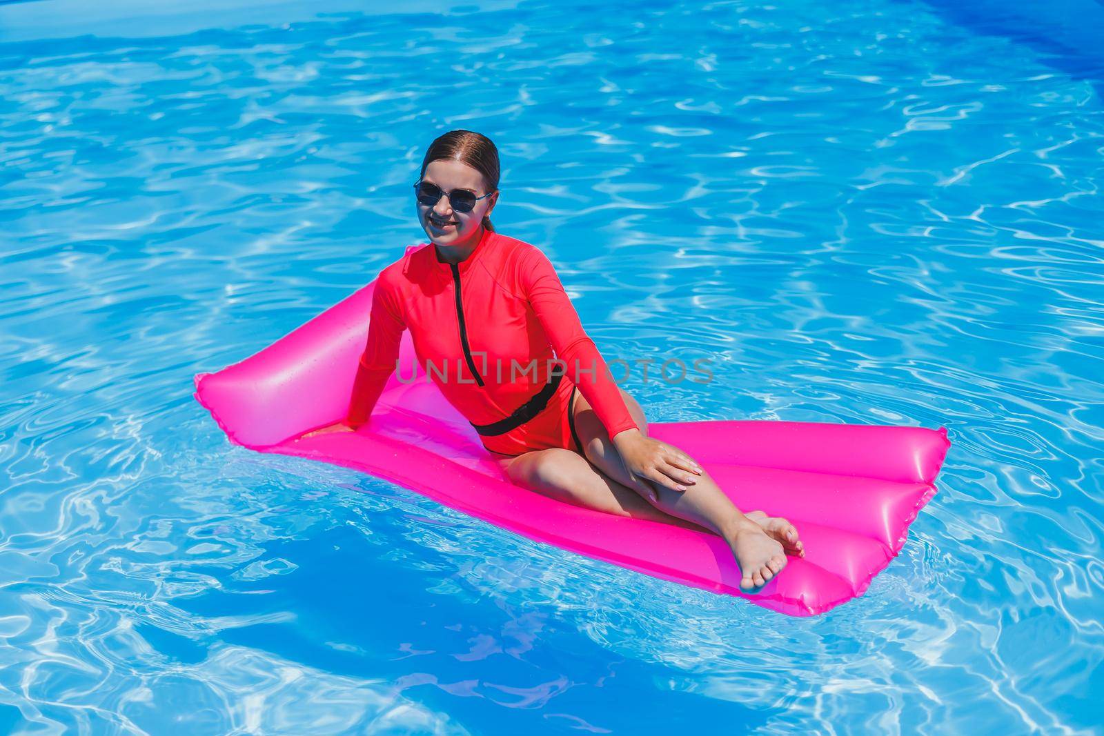 Happy young woman in sunglasses and pink bikini floating on inflatable mattress by the pool. Summer vacation concept. Travel by sea