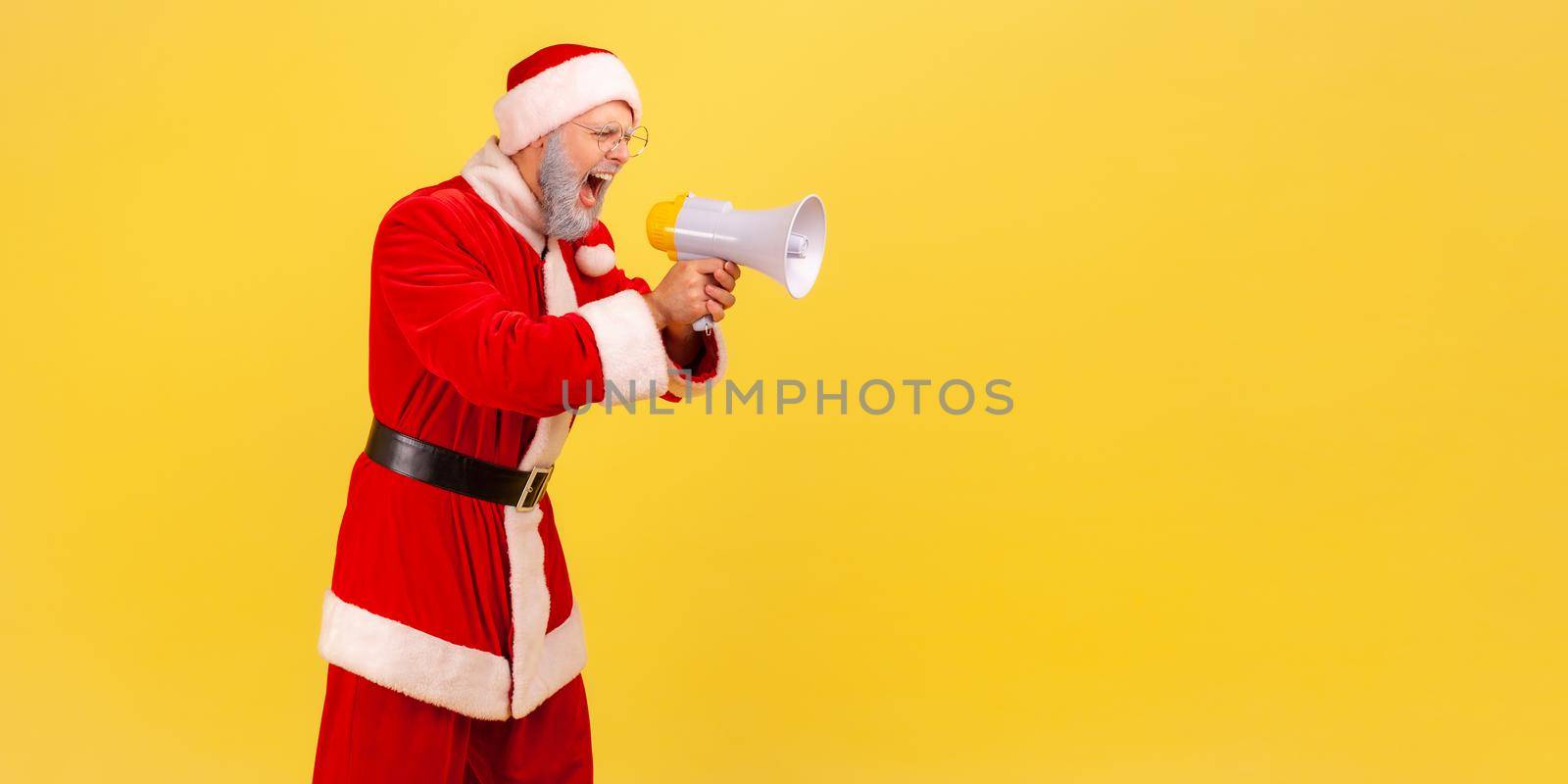 Side view of angry screaming elderly man with gray beard wearing santa claus costume holding megaphone and yelling with aggressive expression. Indoor studio shot isolated on yellow background.