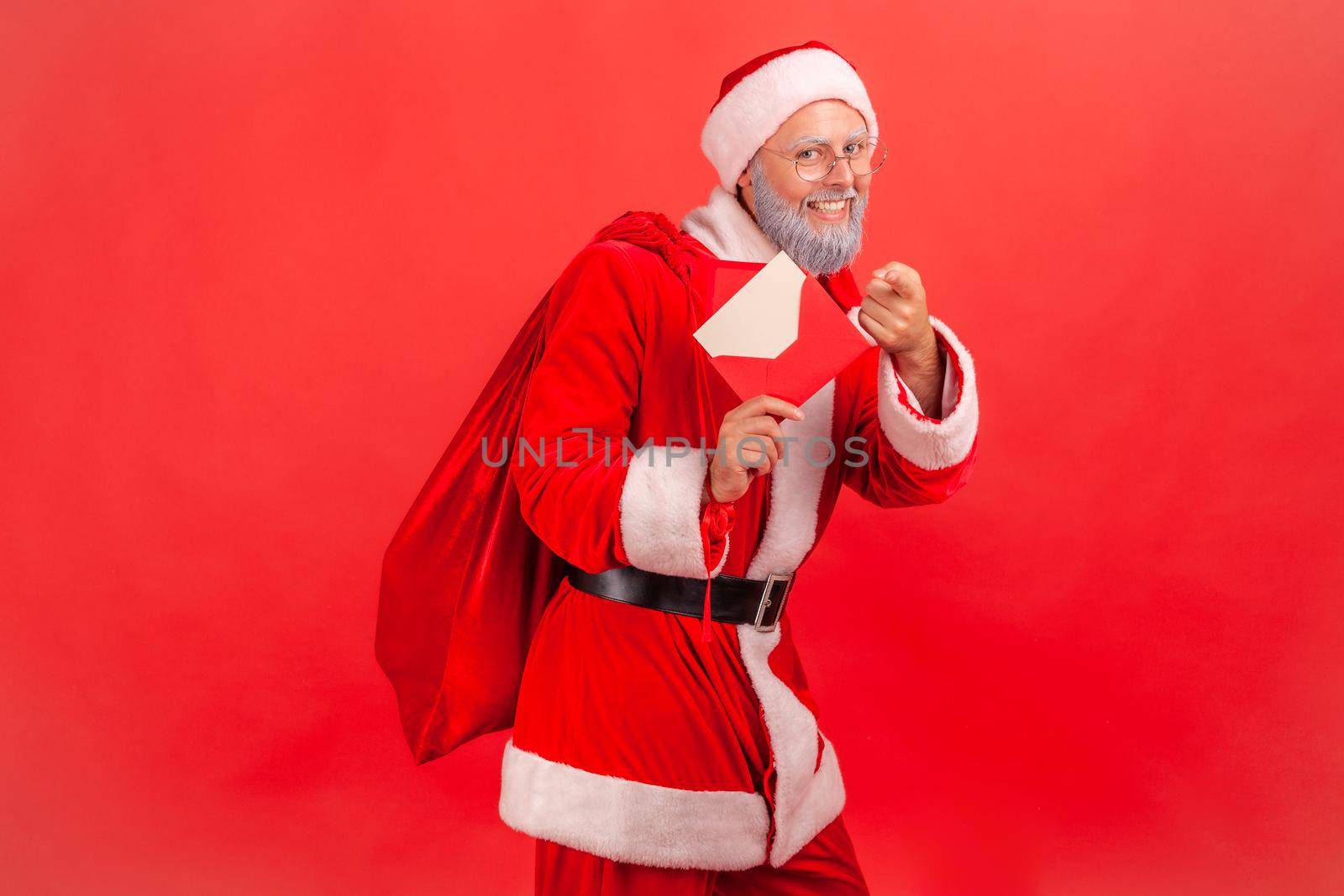 Happy smiling elderly man wearing santa claus costume standing with beg with presents and holding letter with congratulations, pointing to camera. Indoor studio shot isolated on red background.