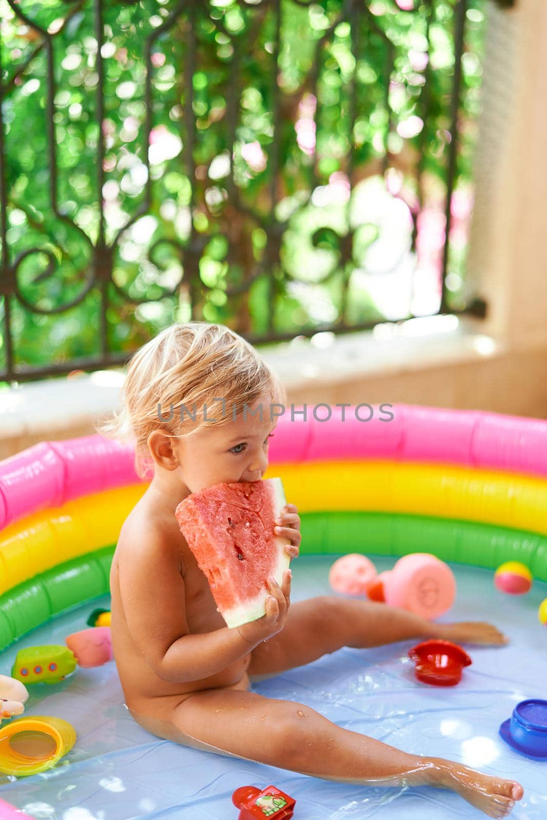 Little girl eats watermelon sitting in an inflatable pool on the balcony by Nadtochiy