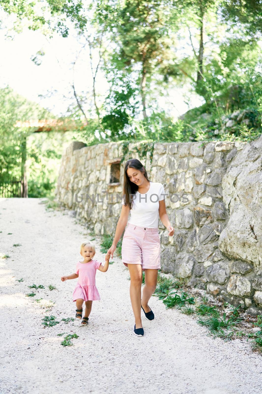 Mom with a little girl walk along the path past the stone wall in the park. High quality photo