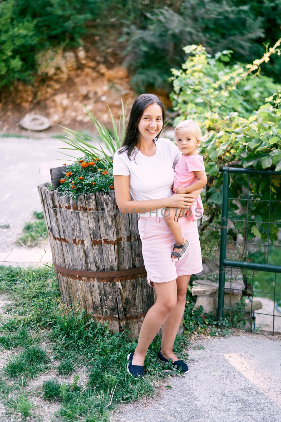Smiling mom with a little daughter in her arms near a wooden barrel with flowers. High quality photo