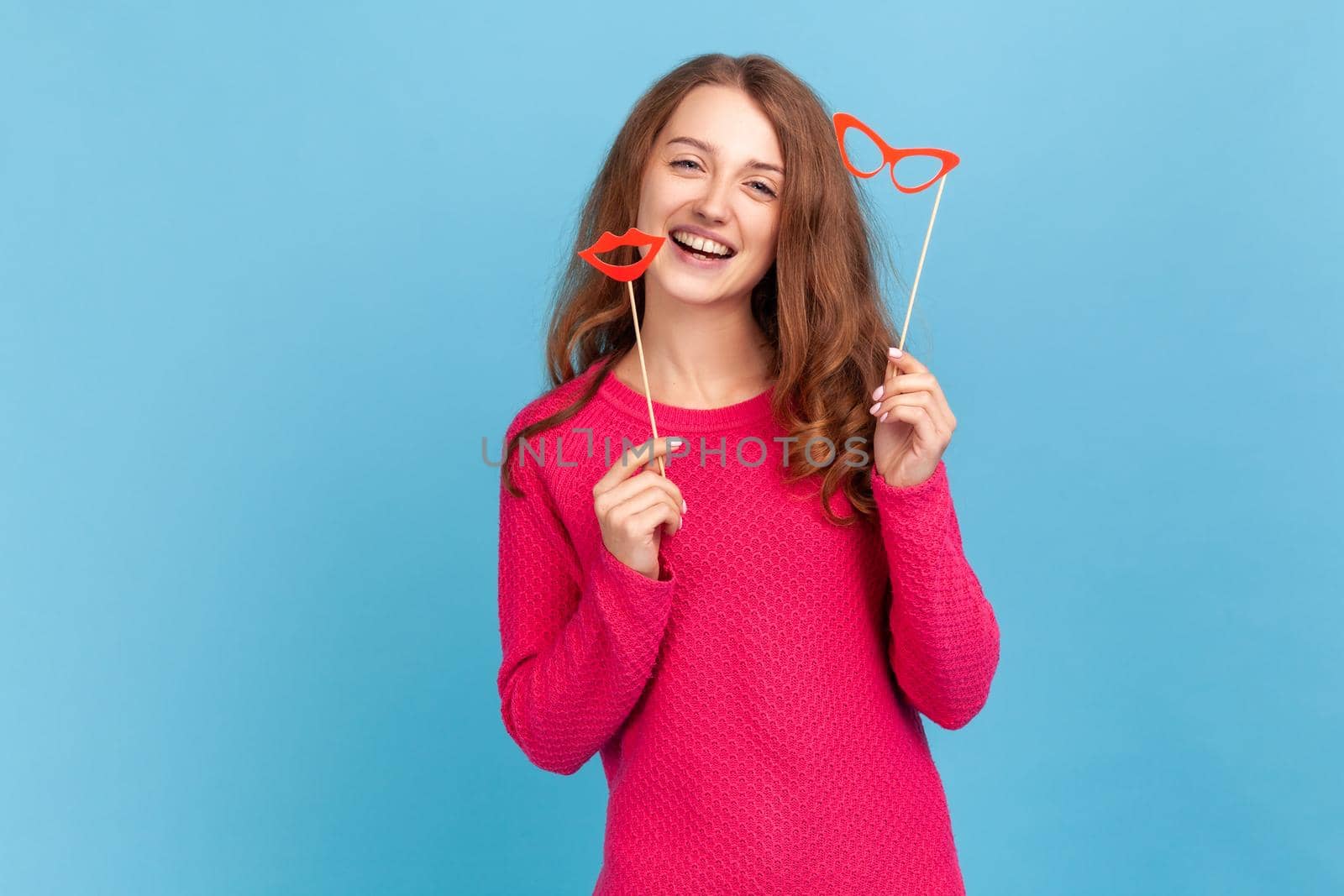 Attractive woman with festive mood, wearing pink pullover, holding party props, celebrating holiday, showing paper lips and glasses. Indoor studio shot isolated on blue background.