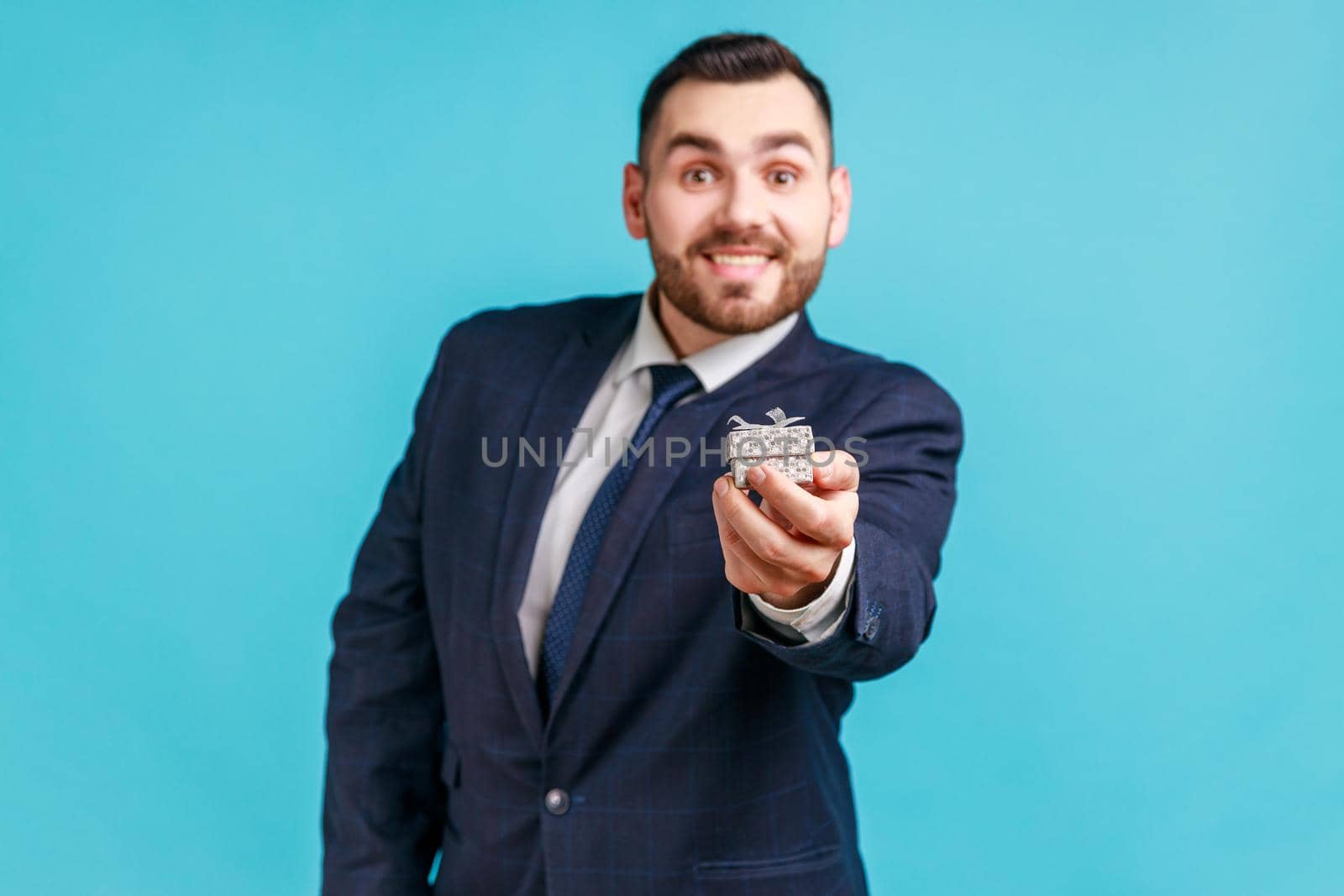 Happy smiling man wearing dark official style suit holding out little box with ring, making proposal to her girlfriend, looking at camera. Indoor studio shot isolated on blue background.
