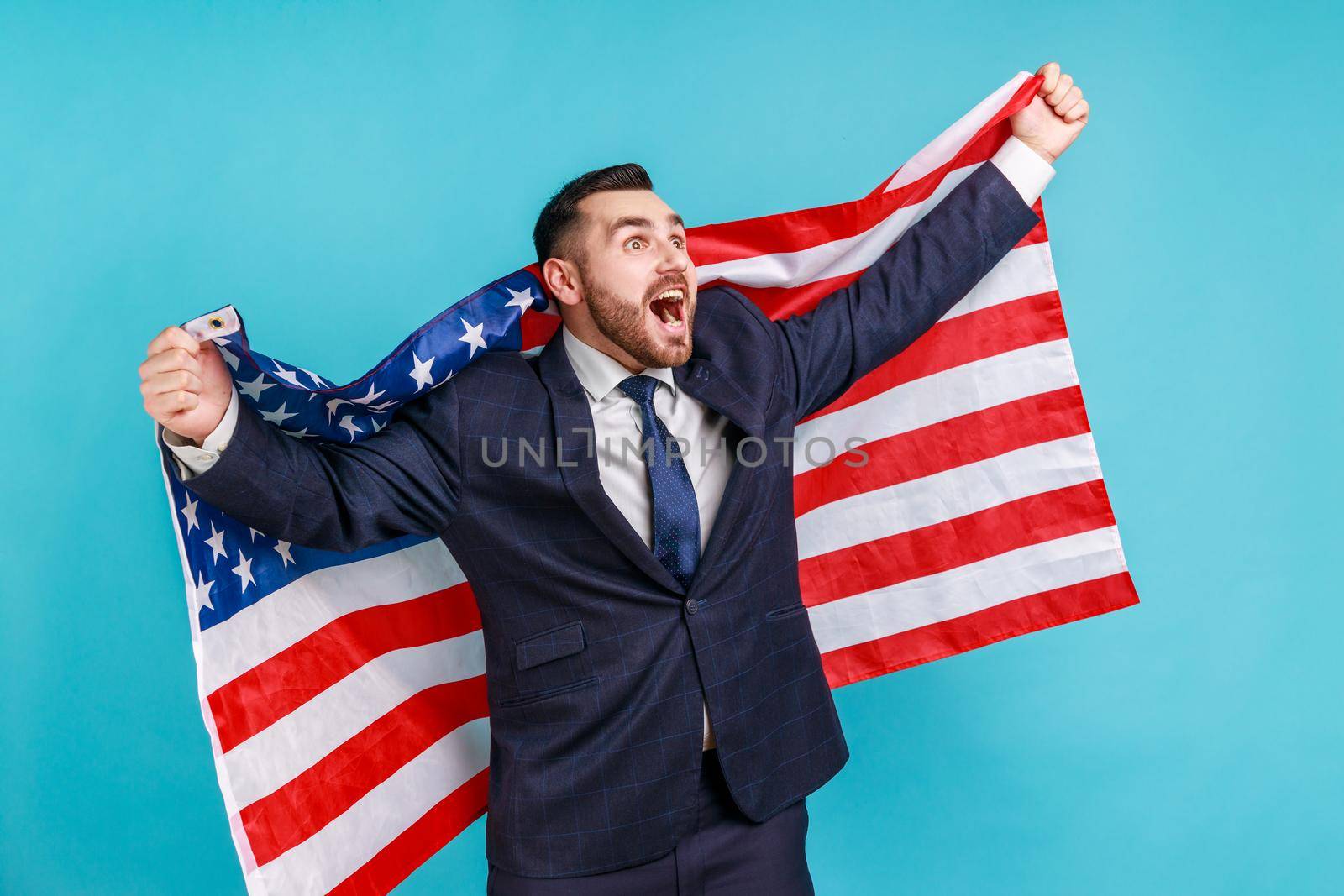 Happy businessman in suit raising American flag, shouting for joy celebrating US Independence day 4th of july, government business support. Indoor studio shot isolated on blue background.