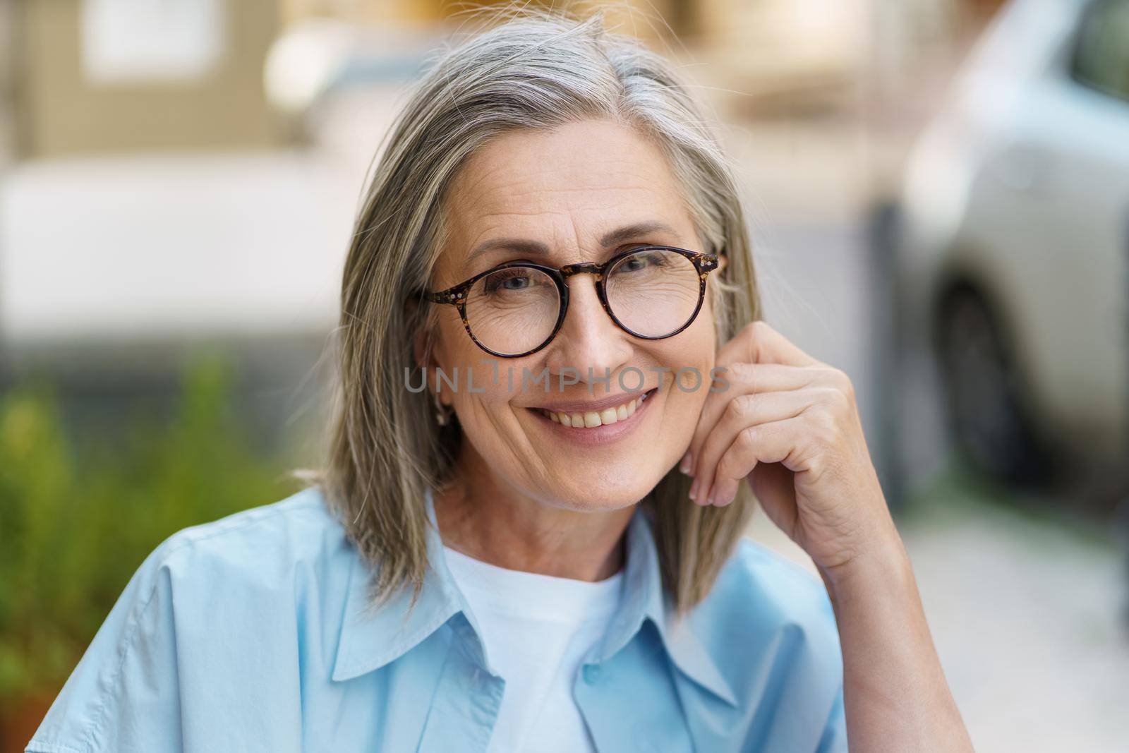 Confident charming mature woman smiling leaned head on hand wearing eye glasses and blue shit sitting outdoors with city background. Portrait of mature woman with grey hair and good skin by LipikStockMedia