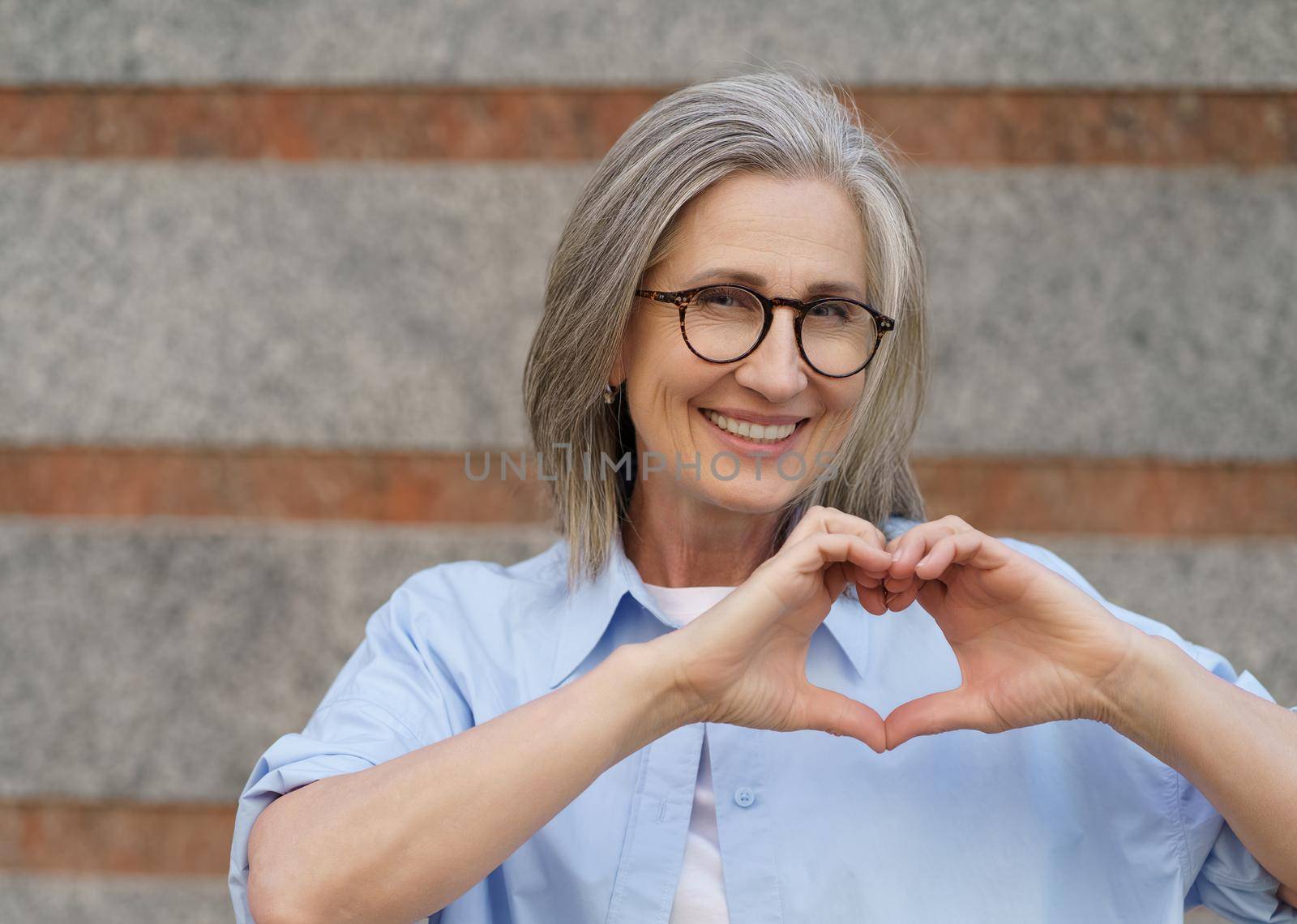 Portrait of attractive mature woman with grey hair showing heart sign gesturing with her hands and smile looking at camera wearing blue casual shirt outdoors. Mature people beauty and care concept.