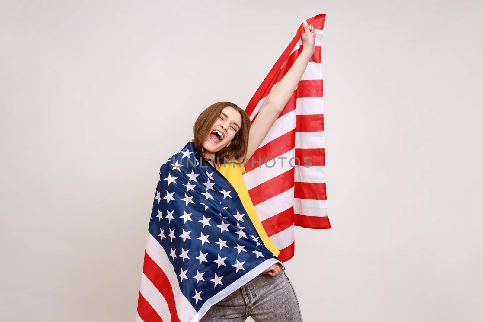 Charming pretty teenager girl with wavy hair, wrapping American flag, yelling happily, raising had up, celebrating Independence day. Indoor studio shot isolated on gray background.