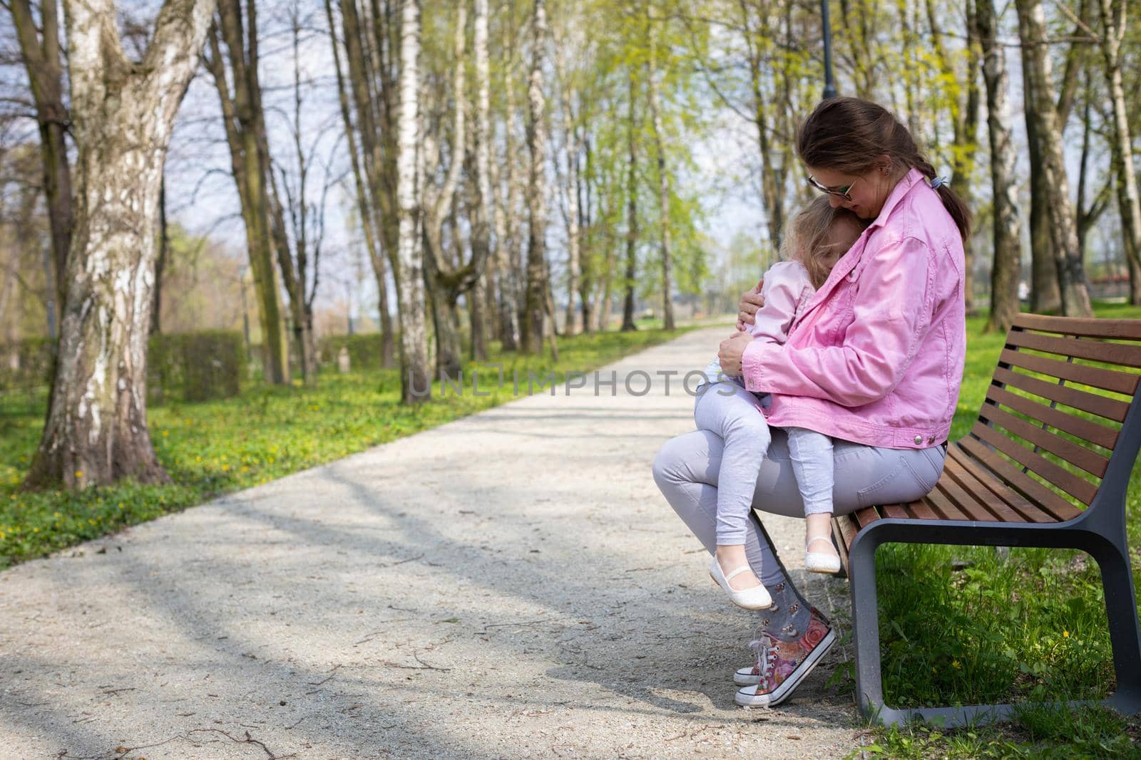 A young mother with her four-year-old daughter are sitting on a bench in a city park. The little girl is cuddling with her mother. A spring sunny day.