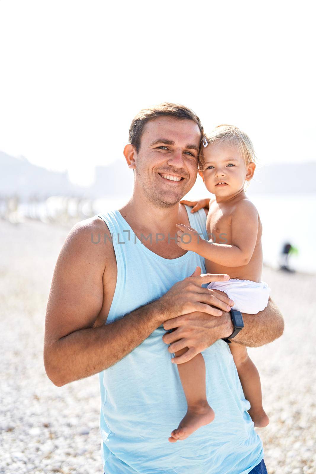 Smiling dad holding little daughter in his arms on the beach. Portrait by Nadtochiy
