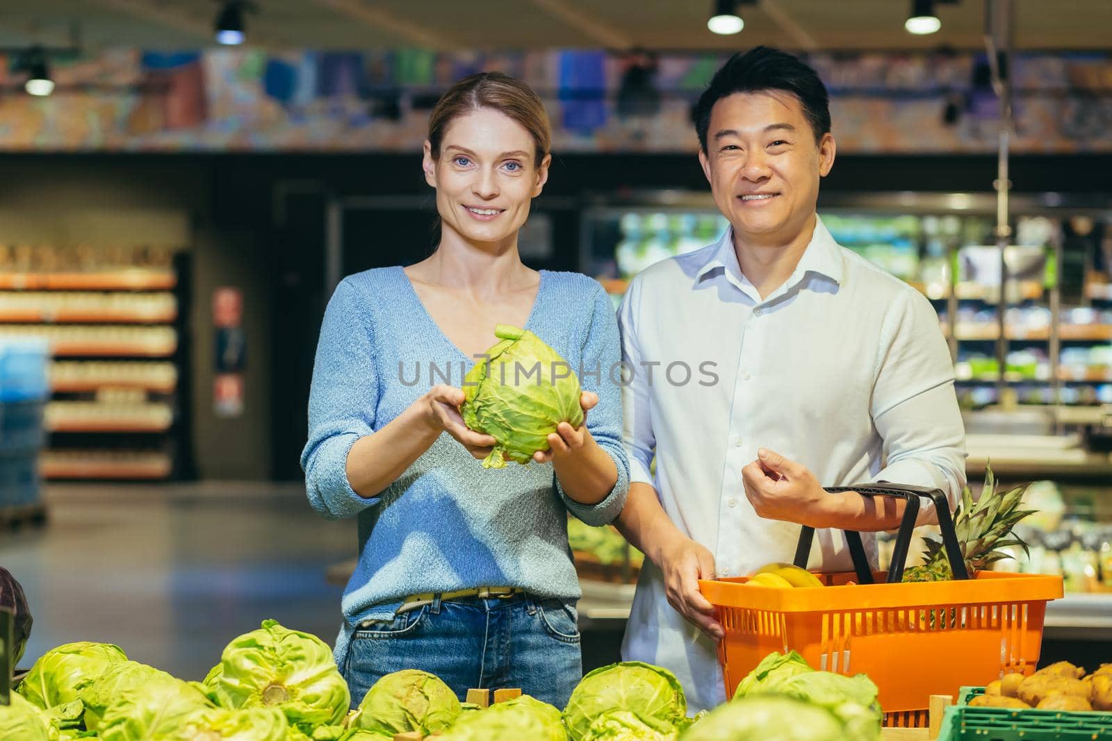 Young married couple, Asian man and blonde woman in supermarket, shoppers choose vegetables in vegetable section, portrait of happy shoppers, smiling looking at camera