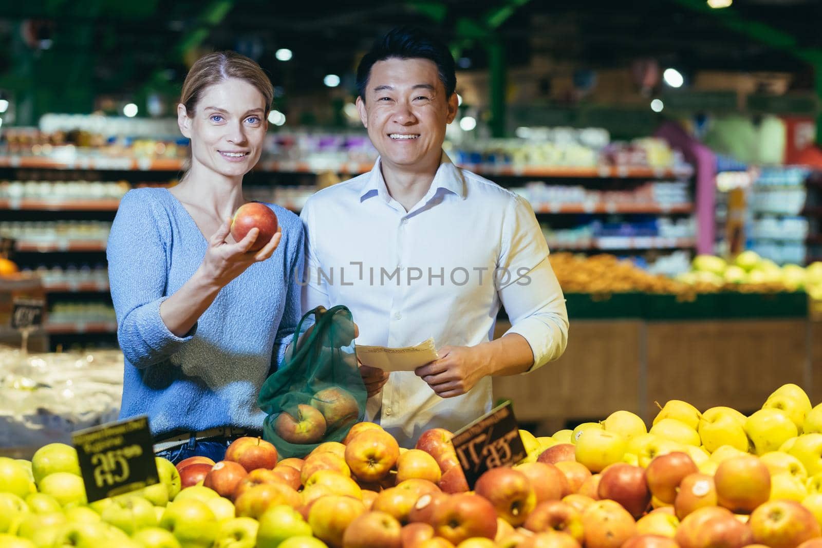 Portrait of young multi-ethnic family, Asian man and woman in supermarket shopping, smiling and looking at camera, couple chooses apple fruit