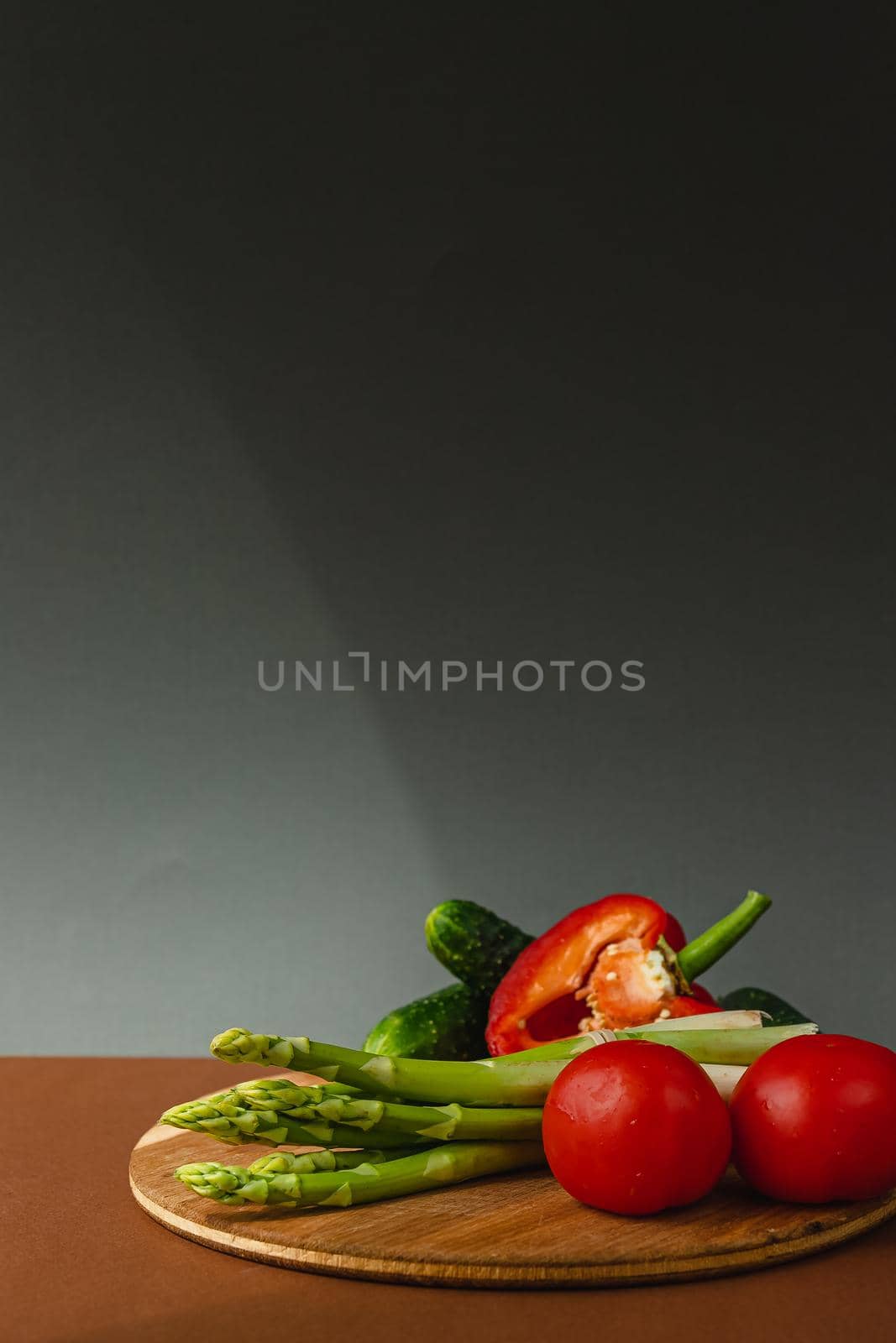 Vegetables lie on a wooden board: tomatoes, asparagus, cucumbers, red bell peppers. brown, dark gray background. place for text