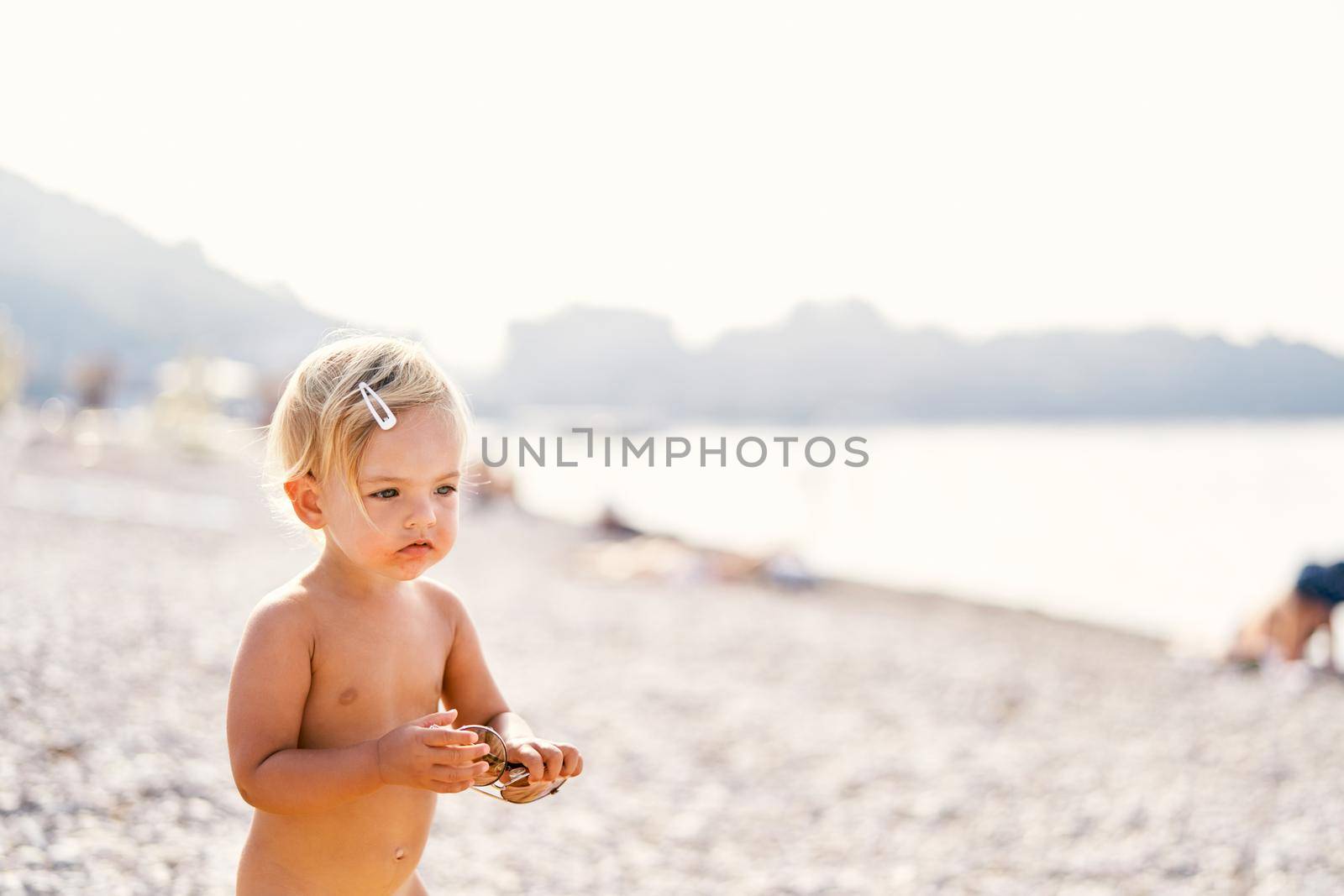Little girl with sunglasses in her hands stands on the beach by Nadtochiy