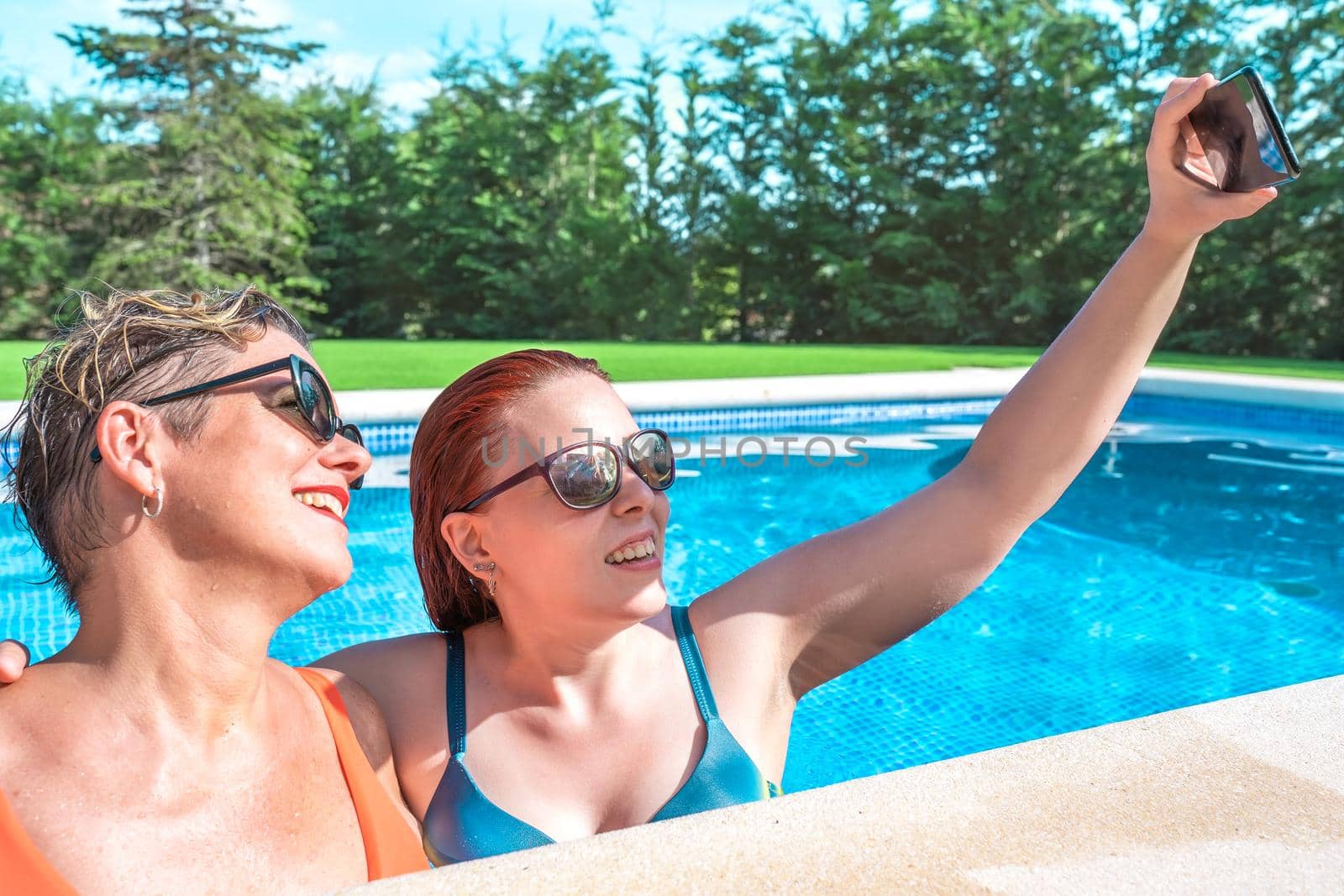 mother and daughter hugging, taking a selfie with their smart phone inside the pool on a sunny day. women enjoying summer holidays together. holiday and travel concept. by CatPhotography