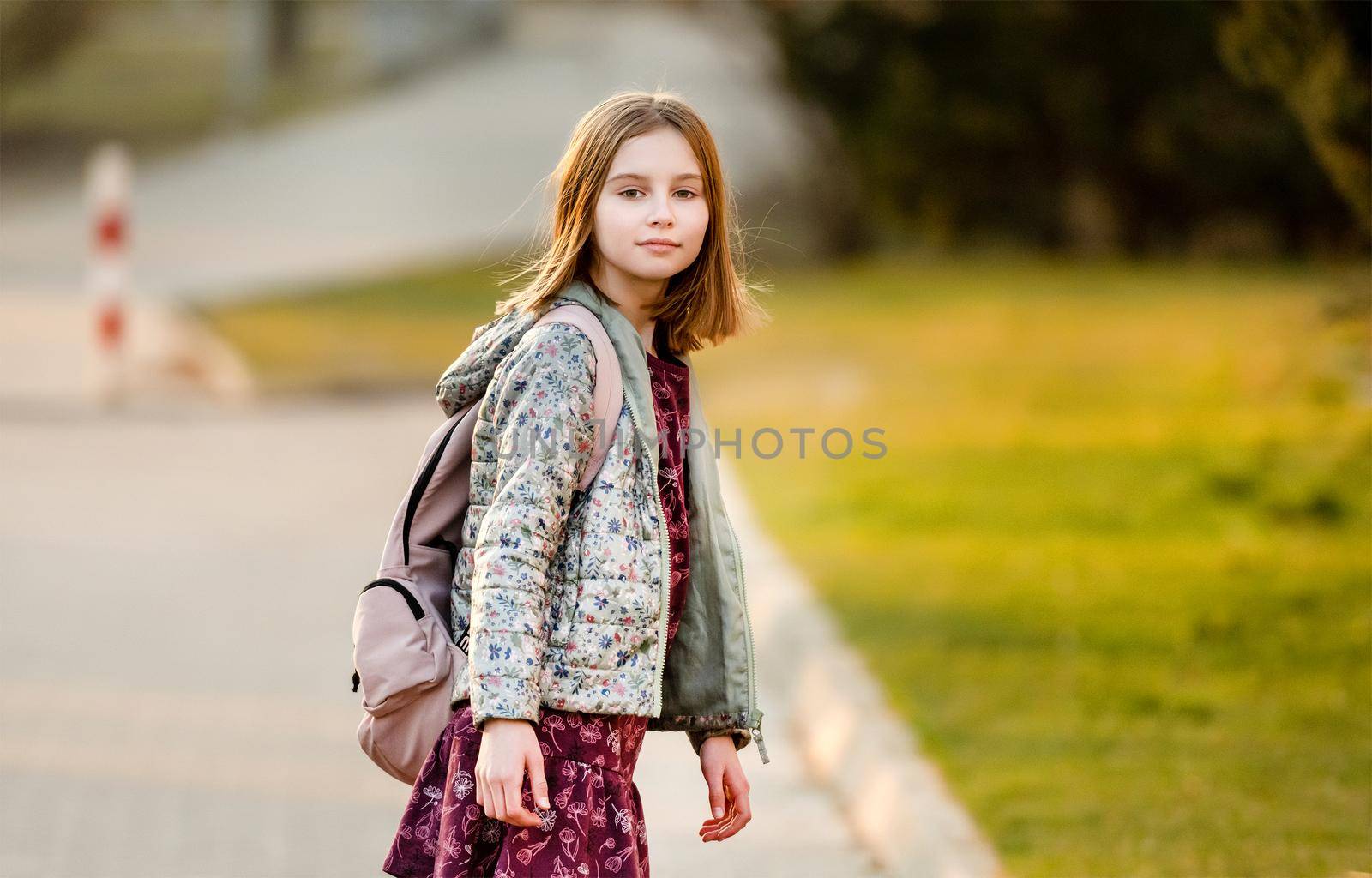 Preteen girl child with backpack at street looking back with beautiful suset light. Pretty schoolgirl going home after class