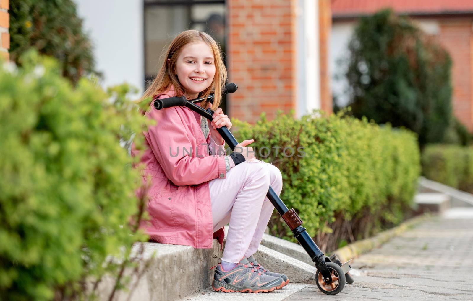 Cute smilling litte girl sitting with a kick scooter on a street near home