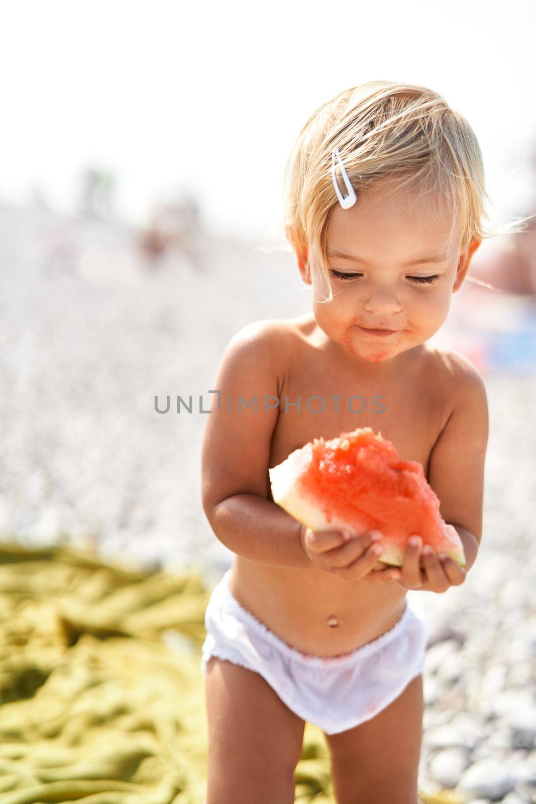 Little smiling girl with a piece of watermelon stands on the beach. High quality photo